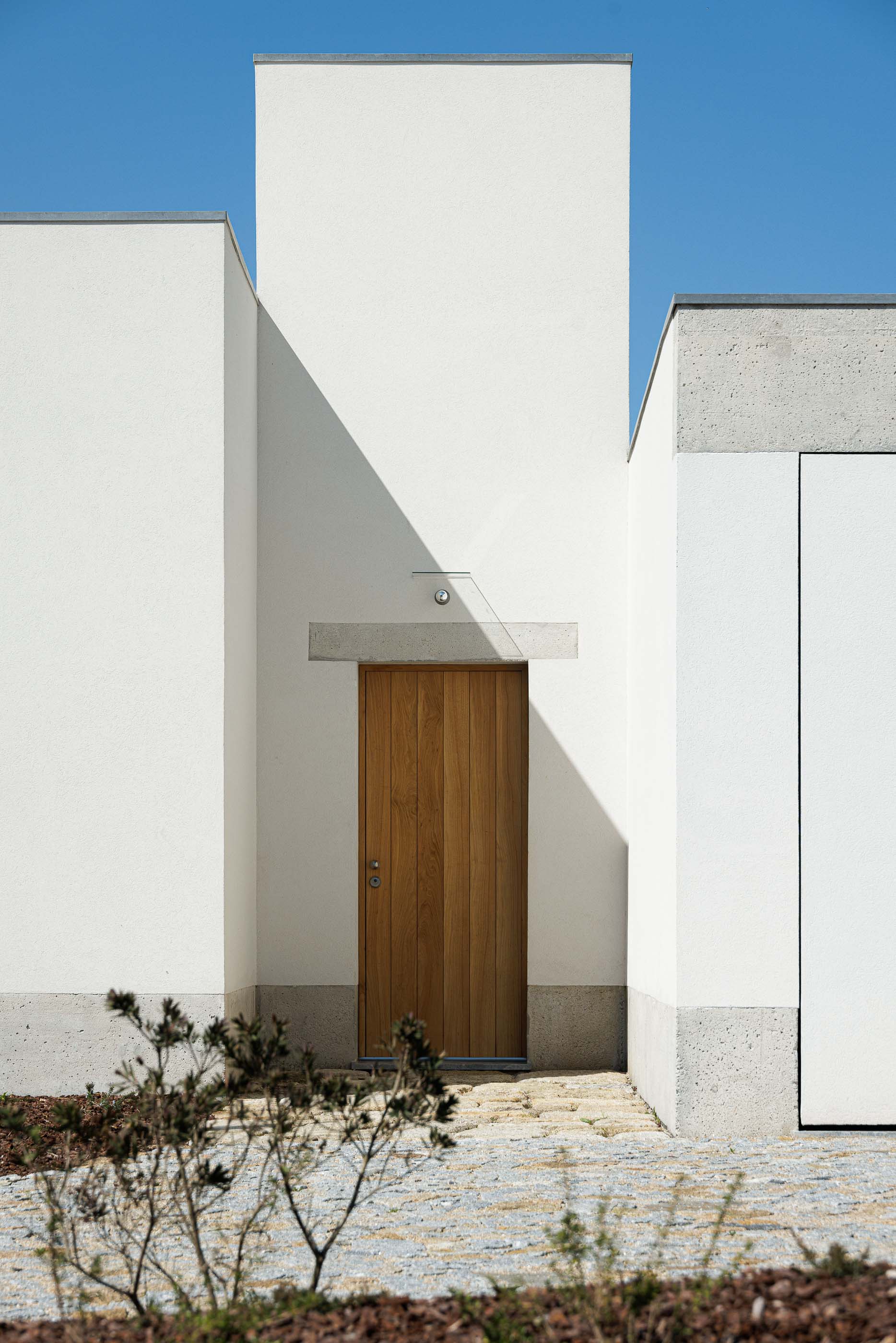 Entrada: Porta de madeira maciça, emoldurada por paredes brancas e detalhes em betão aparente. — Pedrantil House arquitetura Helder da Rocha Arquitectos em Penafiel