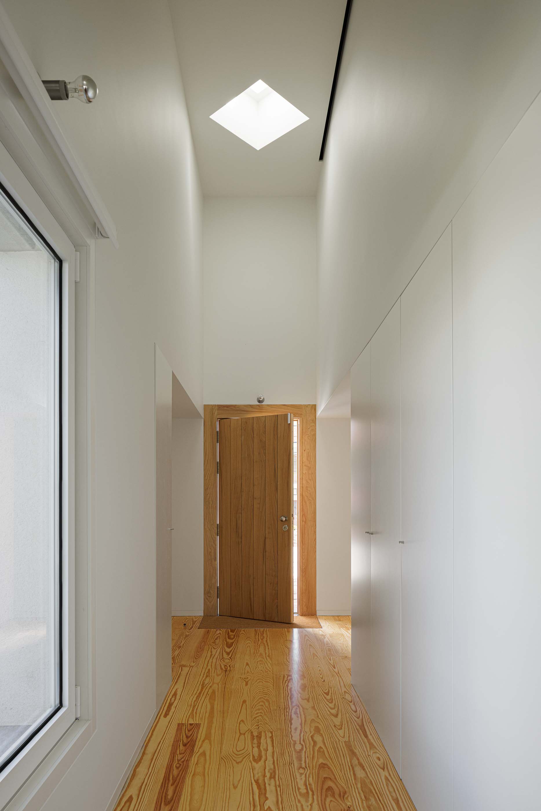 Hall: Corredor com pavimento em madeira, paredes brancas e armários embutidos, iluminado por claraboia. — Pedrantil House arquitetura Helder da Rocha Arquitectos em Penafiel