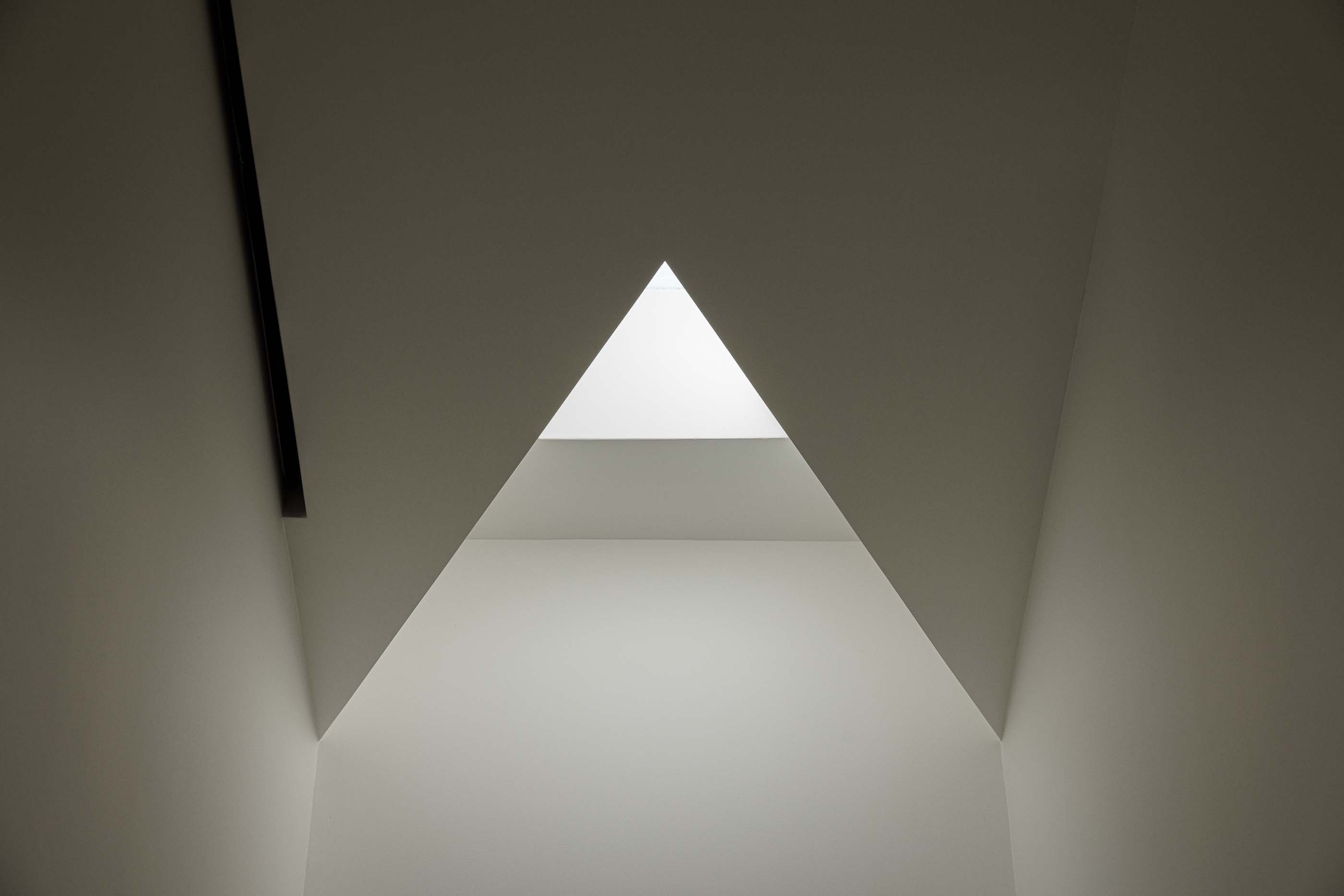 Hall: Claraboia triangular no teto branco, criando um efeito de luz natural dramático. — Pedrantil House arquitetura Helder da Rocha Arquitectos em Penafiel