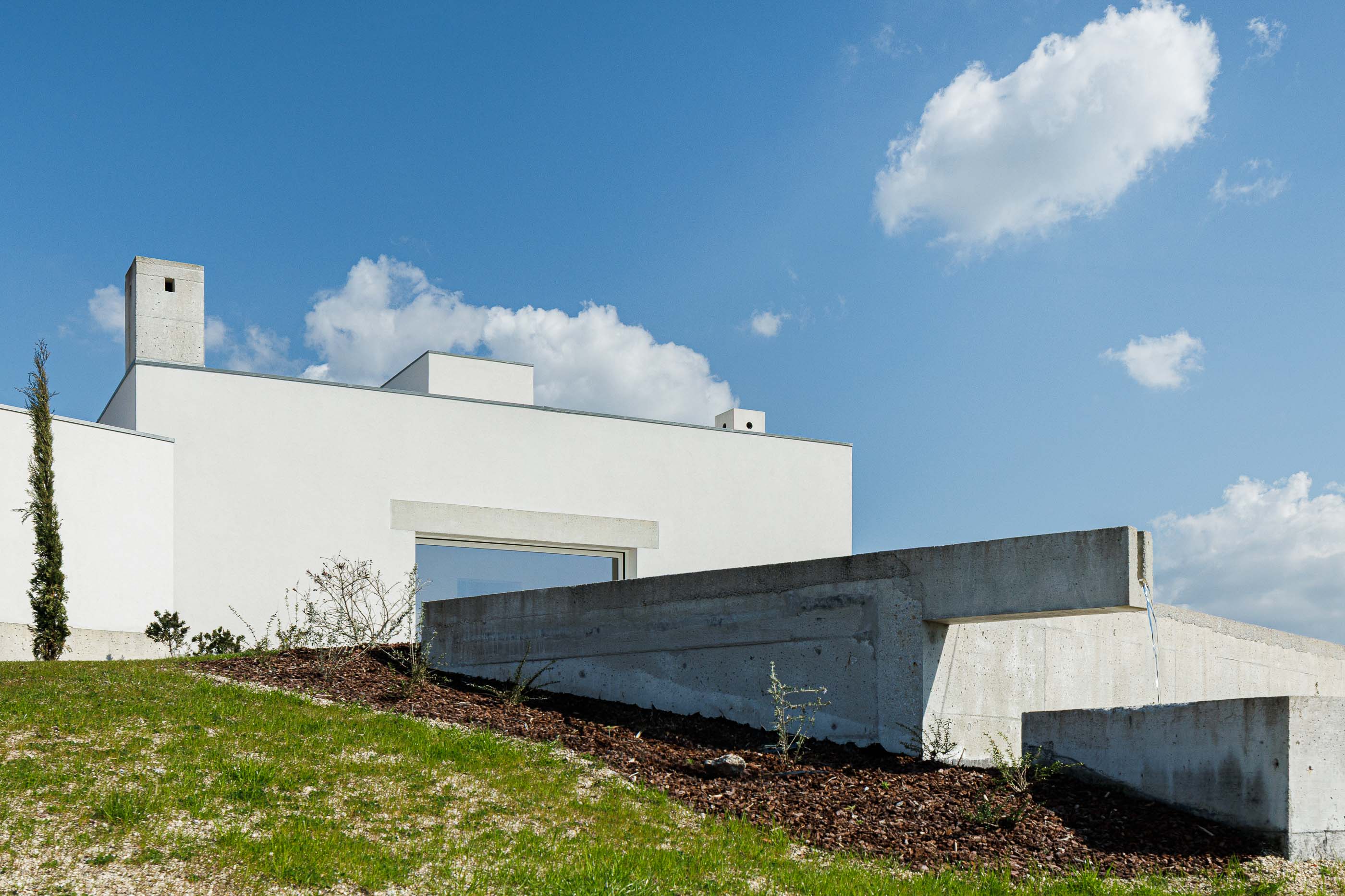 Exterior: Casa branca com elementos em betão, integrada na paisagem natural e com vegetação rasteira. — Pedrantil House arquitetura Helder da Rocha Arquitectos em Penafiel