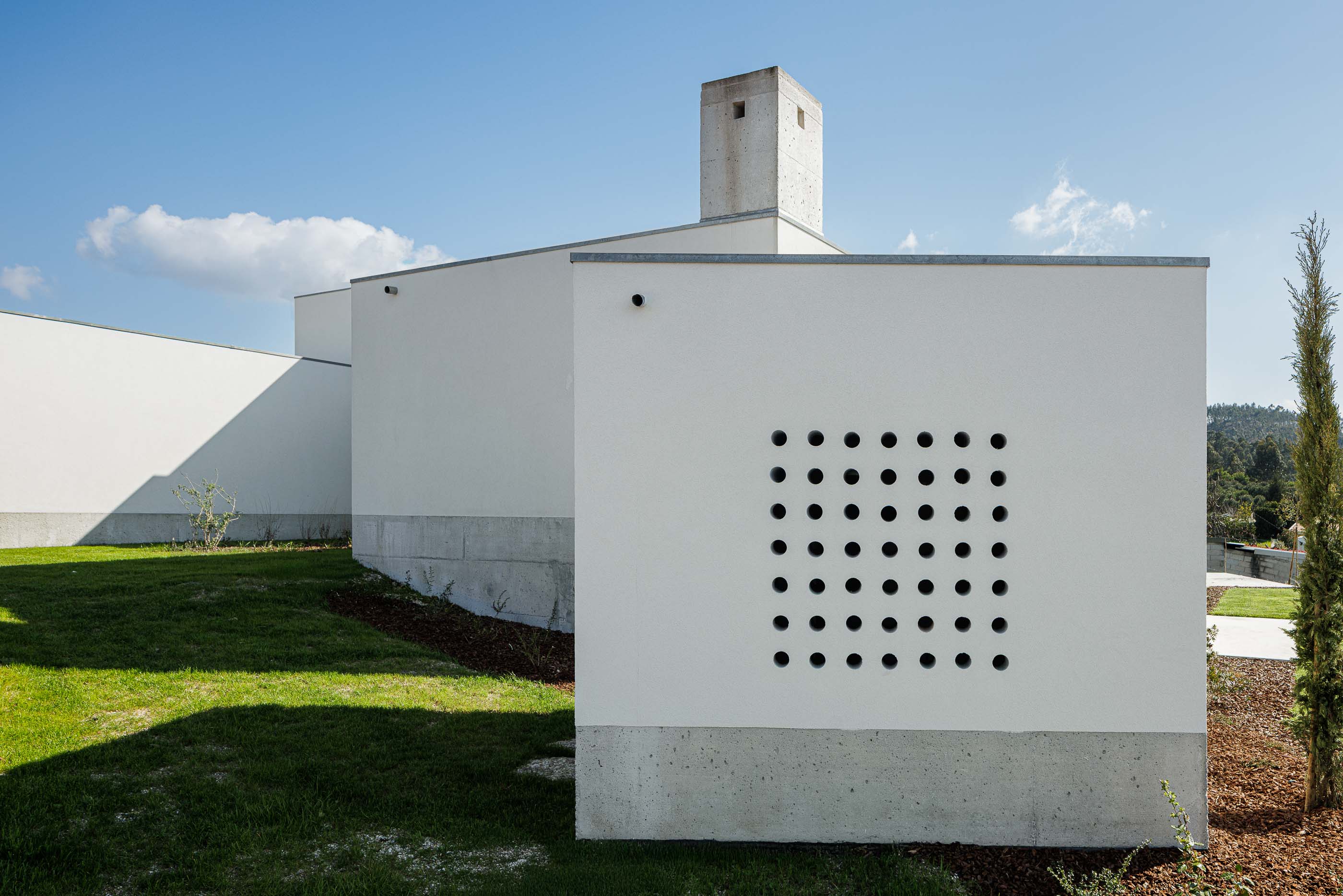 Exterior: Pormenor da fachada com orifícios quadrados, relvado e vegetação, num dia ensolarado. — Pedrantil House arquitetura Helder da Rocha Arquitectos em Penafiel