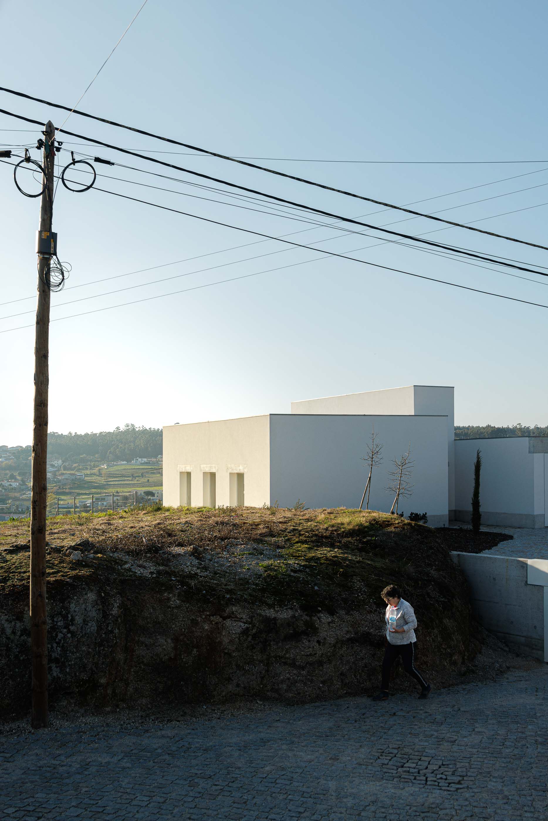 Exterior: Casa branca no topo de um monte, com uma pessoa a caminhar na rua. — Pedrantil House arquitetura Helder da Rocha Arquitectos em Penafiel