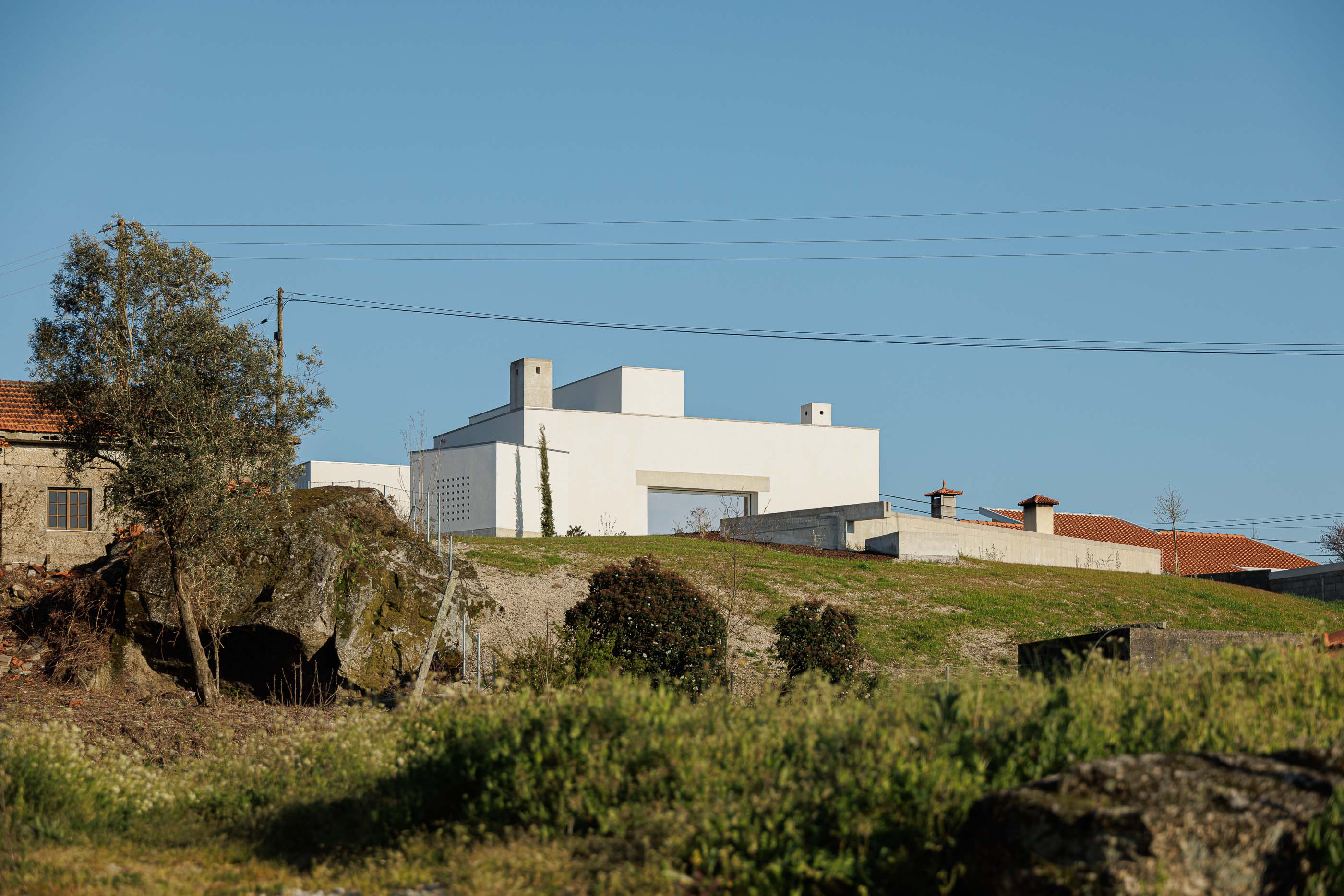 Exterior: Vista da casa a partir de um campo, com vegetação e uma antiga construção em ruínas. — Pedrantil House arquitetura Helder da Rocha Arquitectos em Penafiel