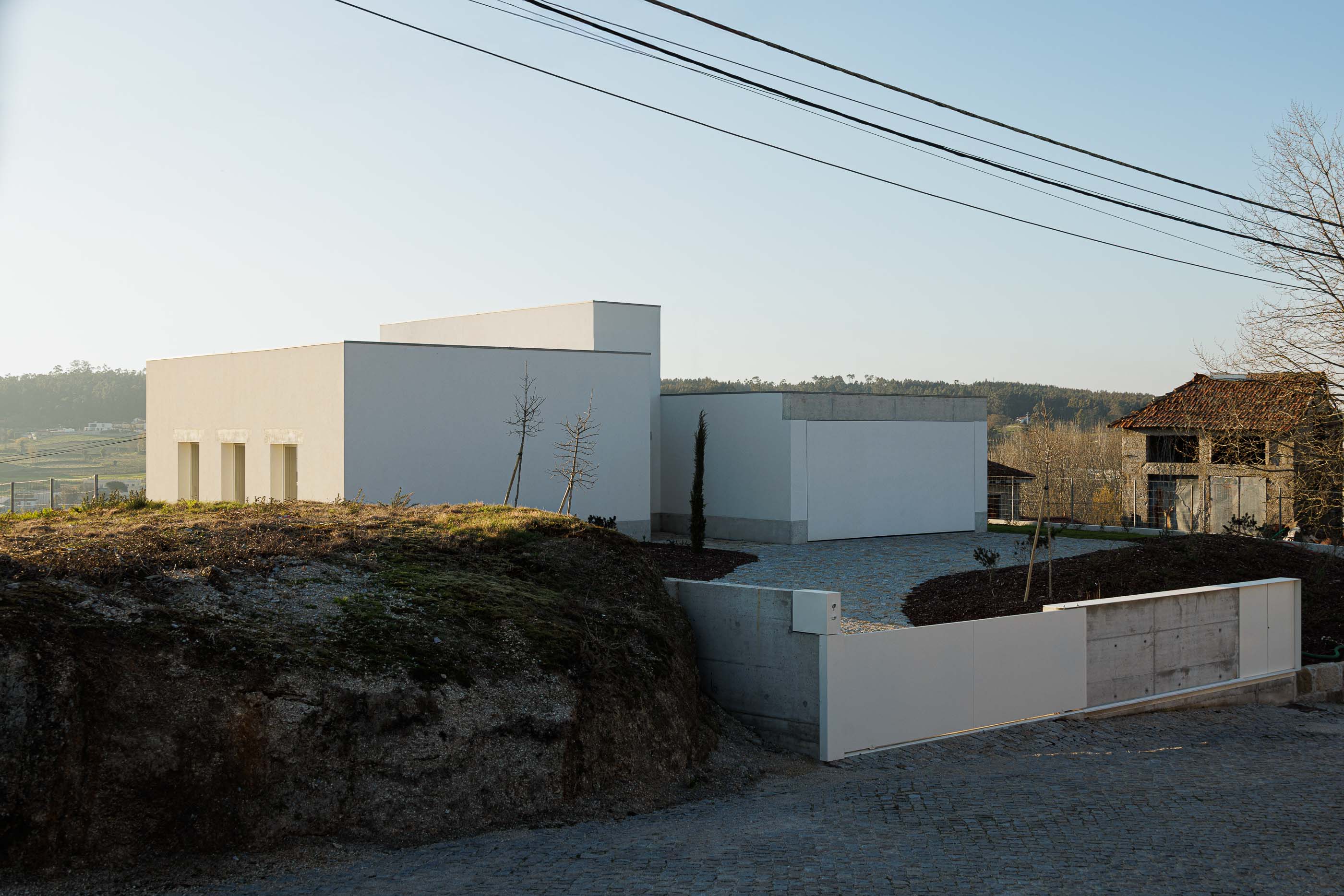 Exterior: Casa branca com volumes geométricos, implantada num terreno rochoso e com vegetação. — Pedrantil House arquitetura Helder da Rocha Arquitectos em Penafiel