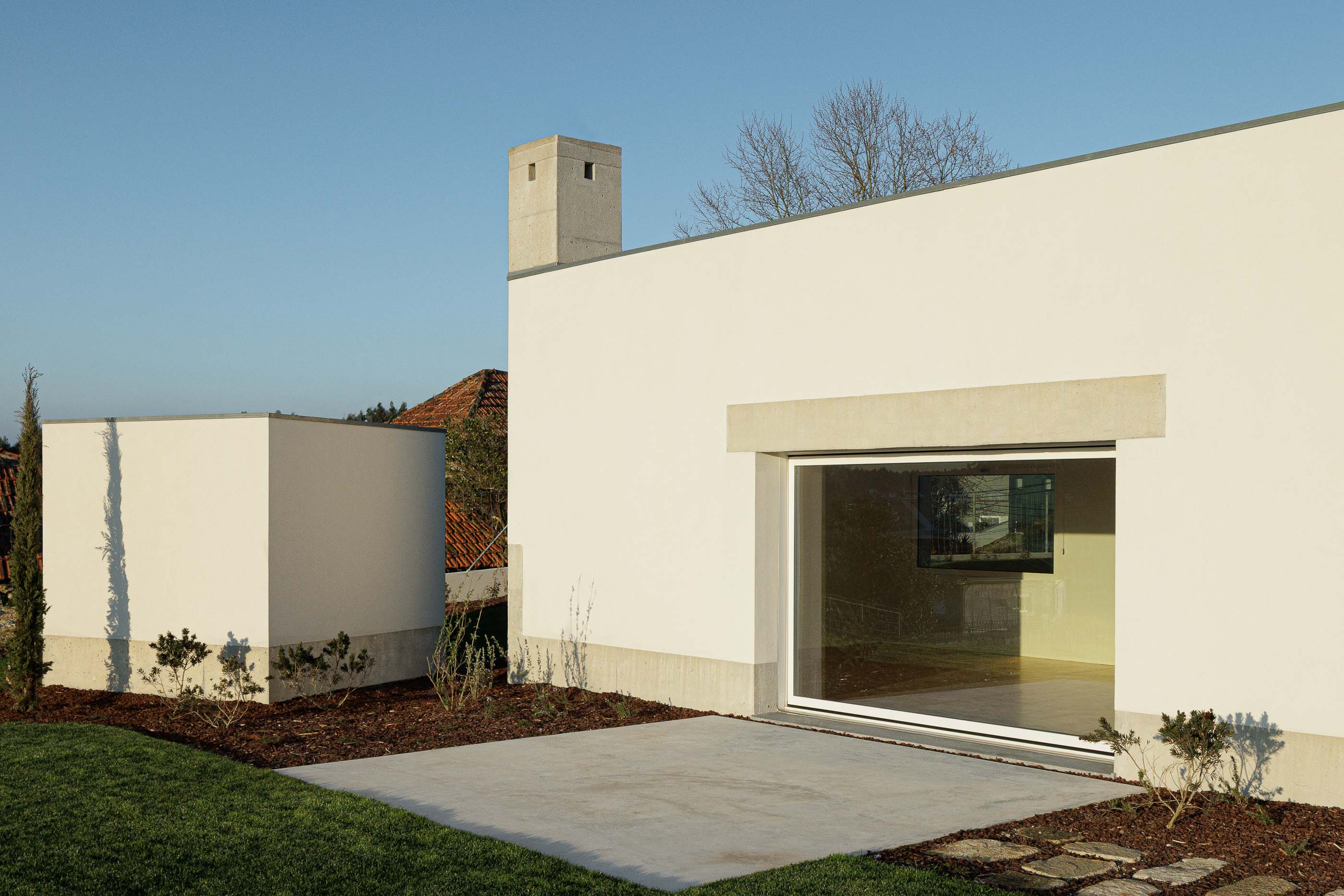 Exterior: Pátio com pavimento em betão e relvado, com a casa ao fundo e vegetação rasteira. — Pedrantil House arquitetura Helder da Rocha Arquitectos em Penafiel