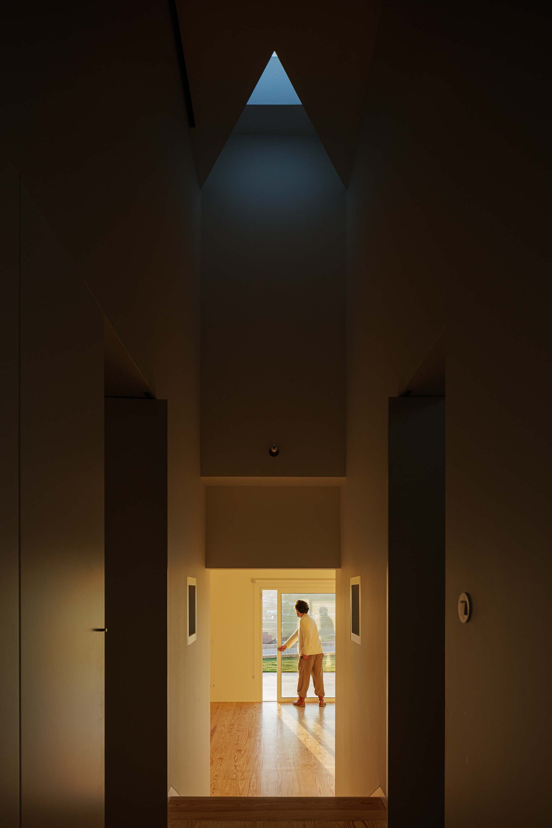 Hall: Corredor com claraboia triangular, pavimento em madeira e uma pessoa a abrir uma porta. — Pedrantil House arquitetura Helder da Rocha Arquitectos em Penafiel