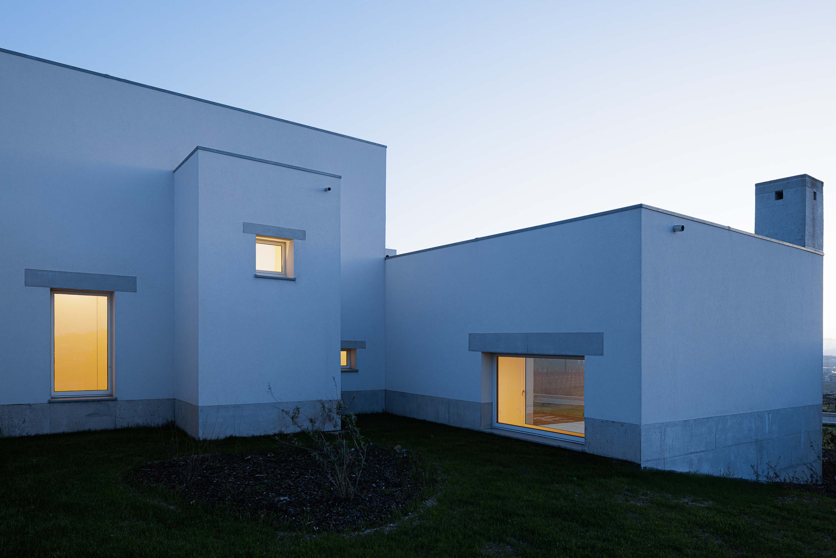 Exterior: Casa branca com janelas iluminadas, vista ao entardecer, com vegetação rasteira. — Pedrantil House arquitetura Helder da Rocha Arquitectos em Penafiel