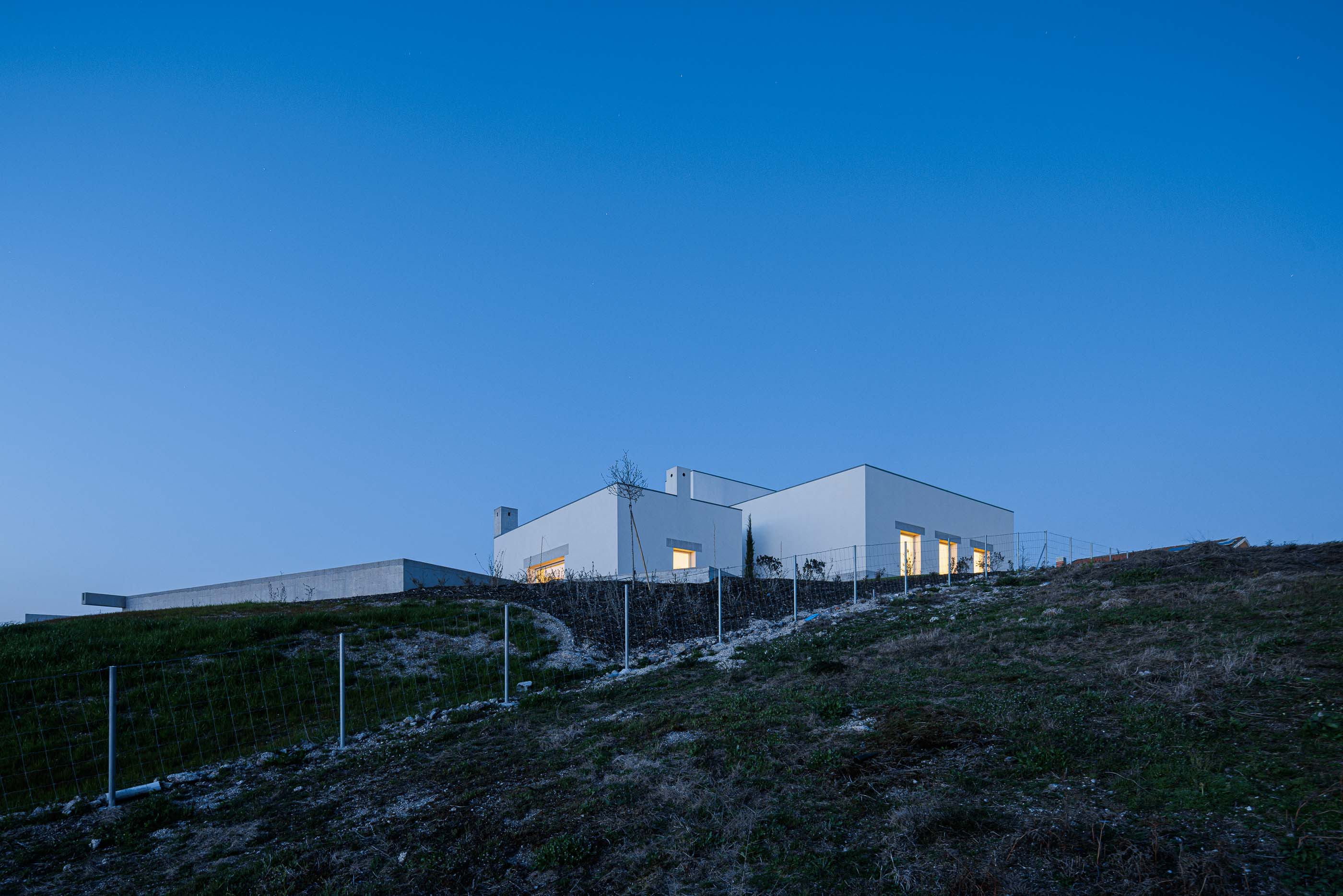 Exterior: Casa branca vista ao anoitecer, com iluminação interior e exterior. — Pedrantil House arquitetura Helder da Rocha Arquitectos em Penafiel