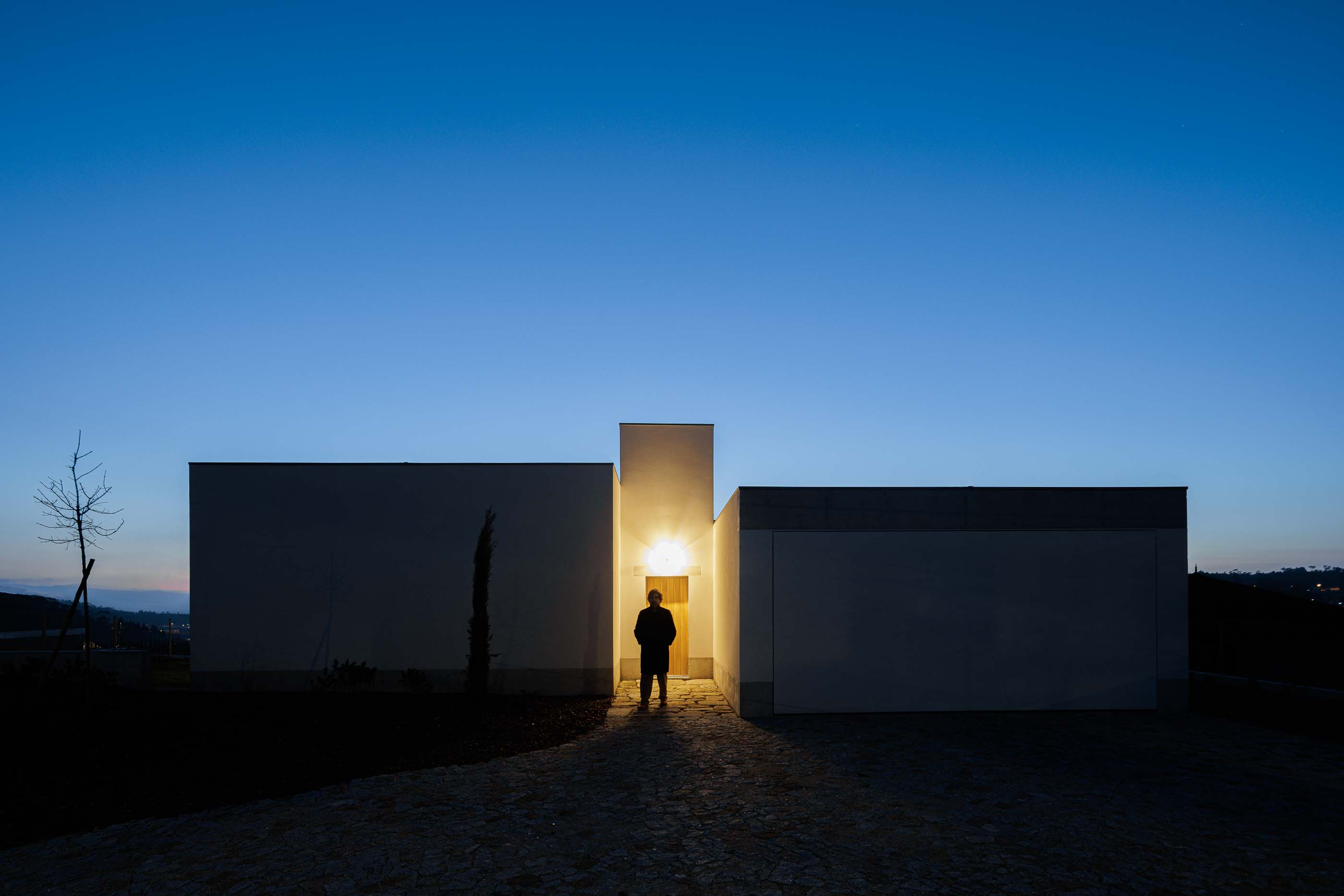 Exterior: Casa branca iluminada ao anoitecer, com uma pessoa à porta. — Pedrantil House arquitetura Helder da Rocha Arquitectos em Penafiel
