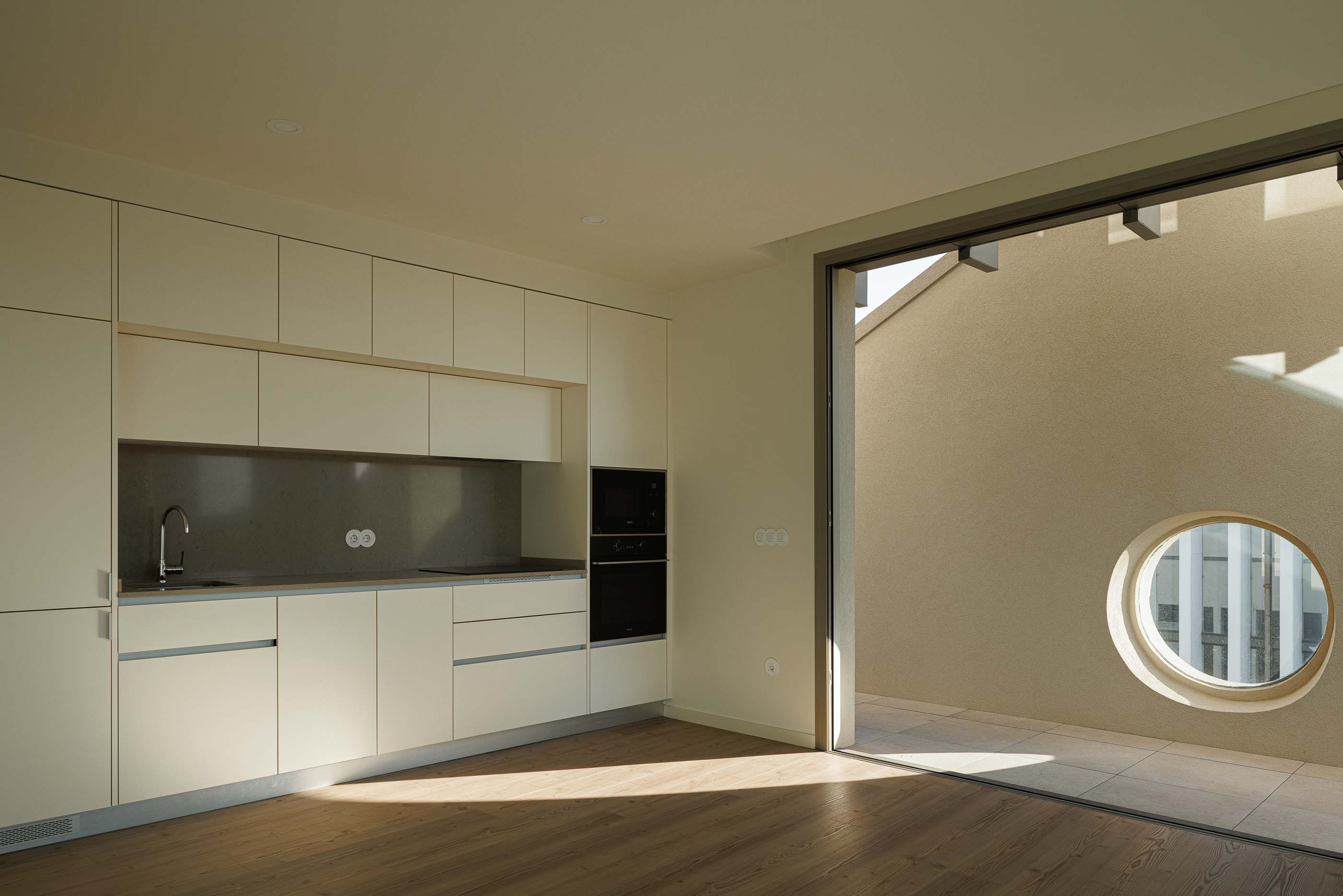 Cozinha moderna com armários brancos, bancada cinza e acesso ao terraço com janela circular. — Edifício Estação arquitetura Sónia Cruz Arquitectura em Aveiro
