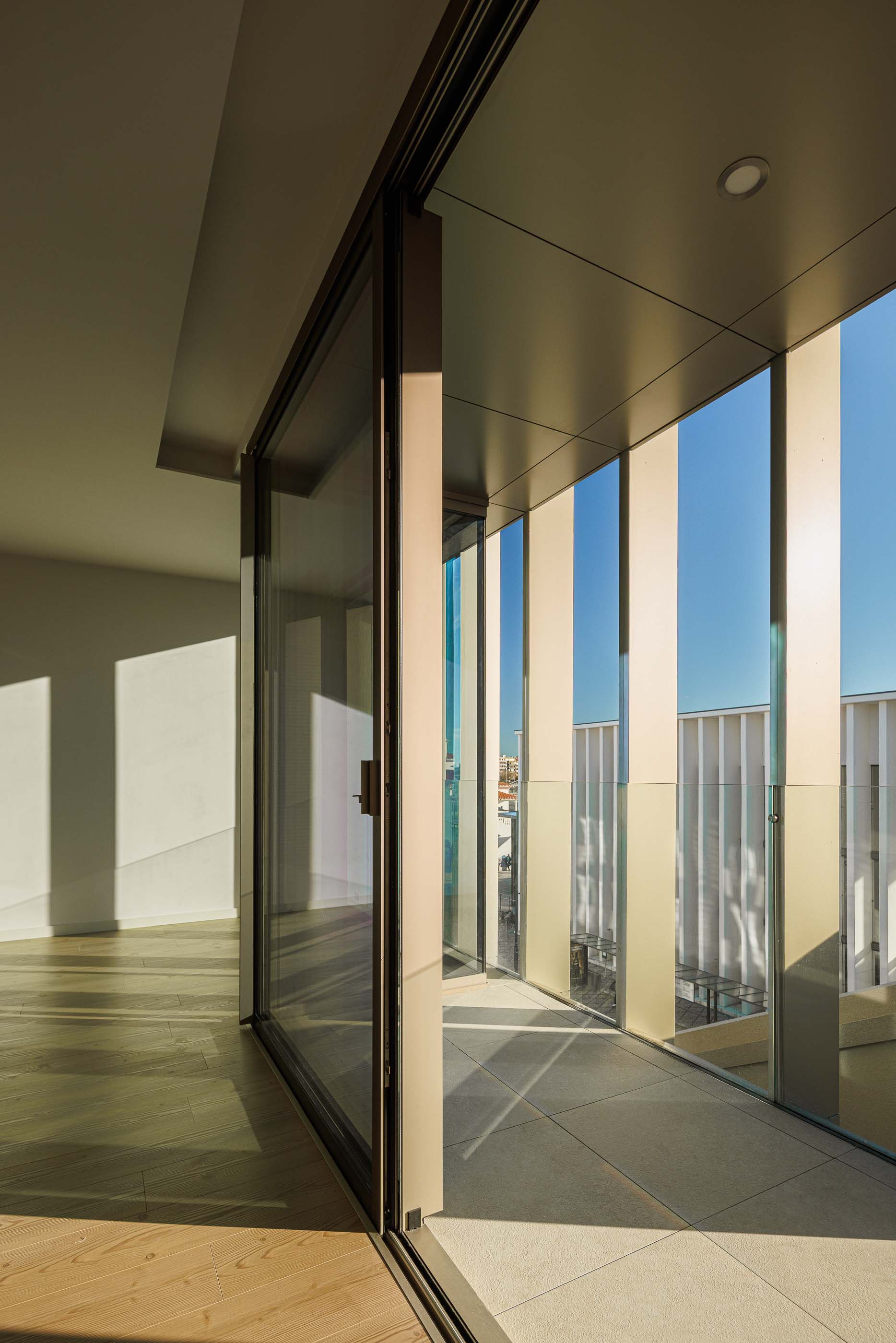 Varanda com pavimento cerâmico, guarda-corpo de vidro e vista para a cidade através das lâminas verticais. — Edifício Estação arquitetura Sónia Cruz Arquitectura em Aveiro