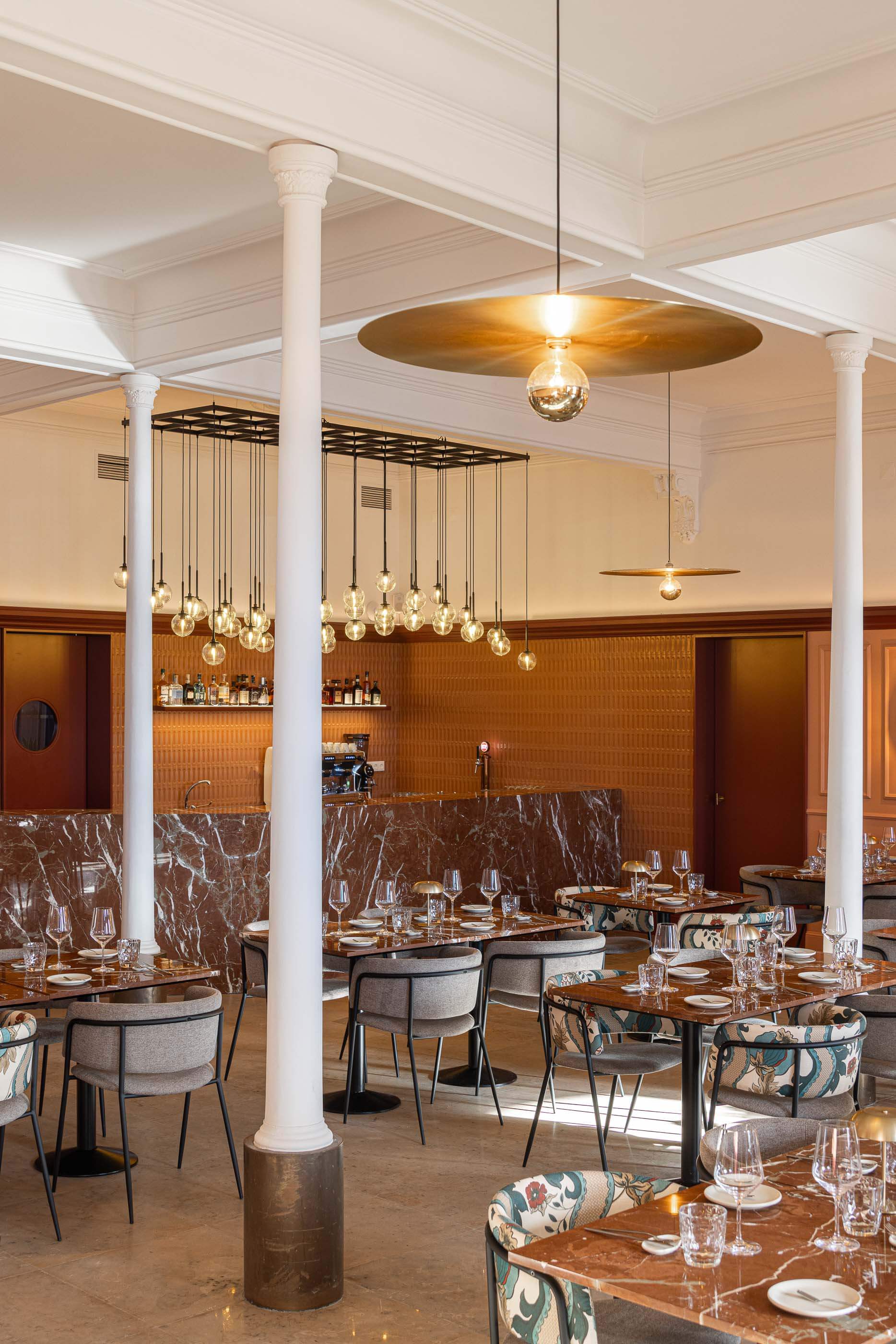 Restaurante: Perspectiva do salão com mesas de mármore, cadeiras cinzentas e coloridas, e bar ao fundo com iluminação suspensa. — Restaurante Concerto arquitetura MiMool Arquitectura & Interiores em vila do conde