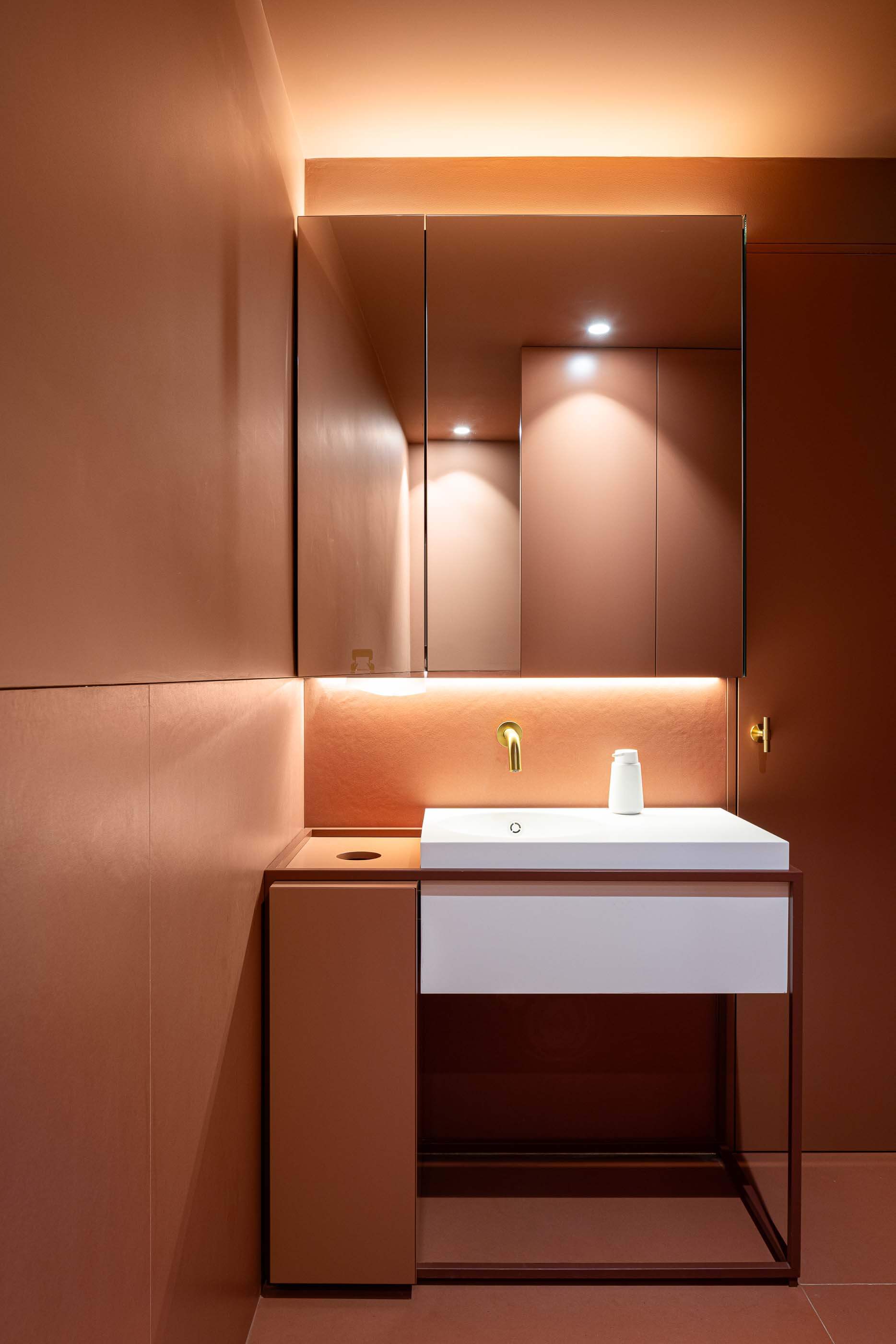 Casa de banho: Lavatório branco com espelho iluminado, torneira dourada e pavimento em tons de terracota. — Restaurante Concerto arquitetura MiMool Arquitectura & Interiores em vila do conde