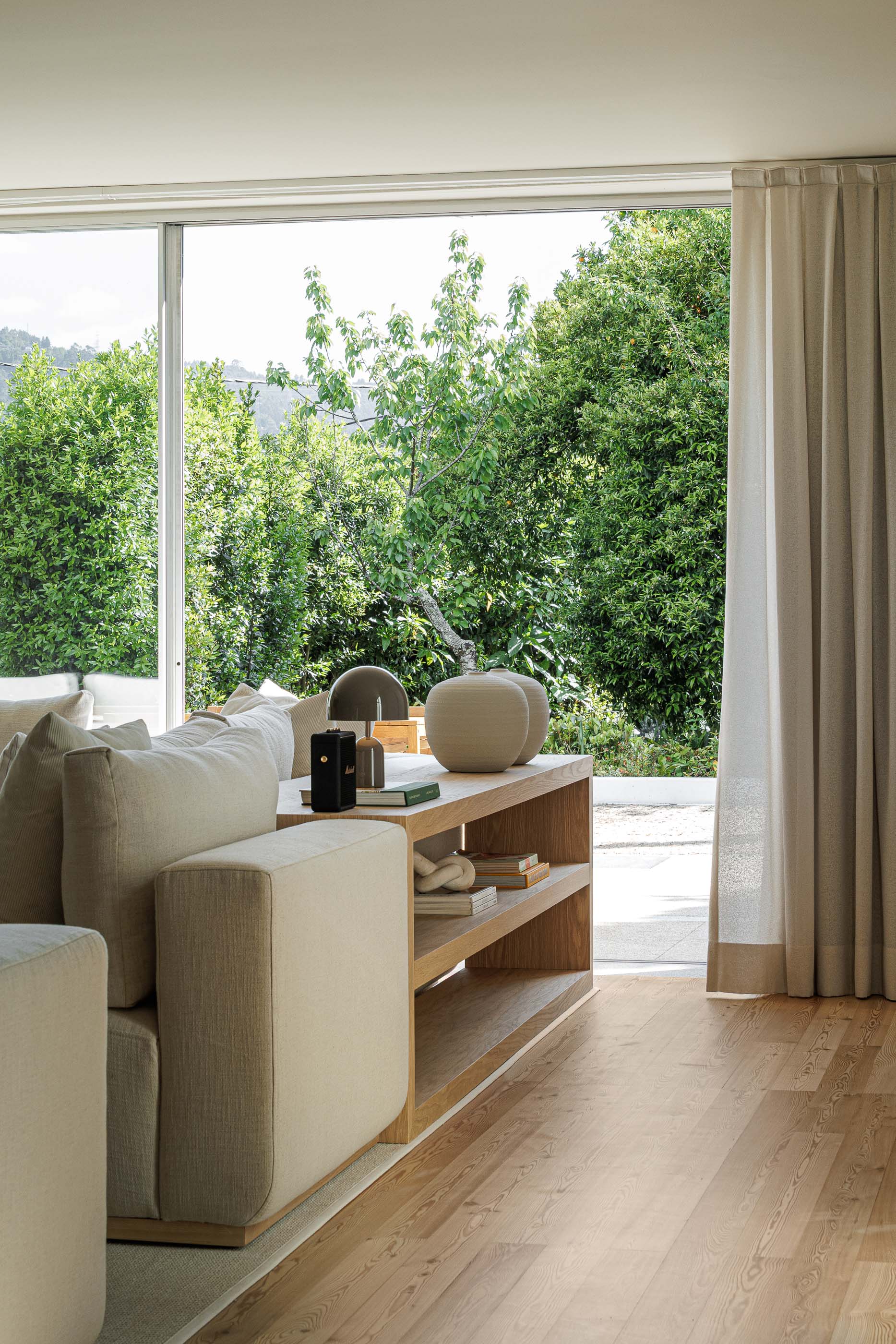 Sala de estar: Sofá branco com almofadas, mesa lateral de madeira e vista para o exterior através de janelas amplas. — Casa no Gerês arquitetura MAT Arquitetura e Interiores em Gerês