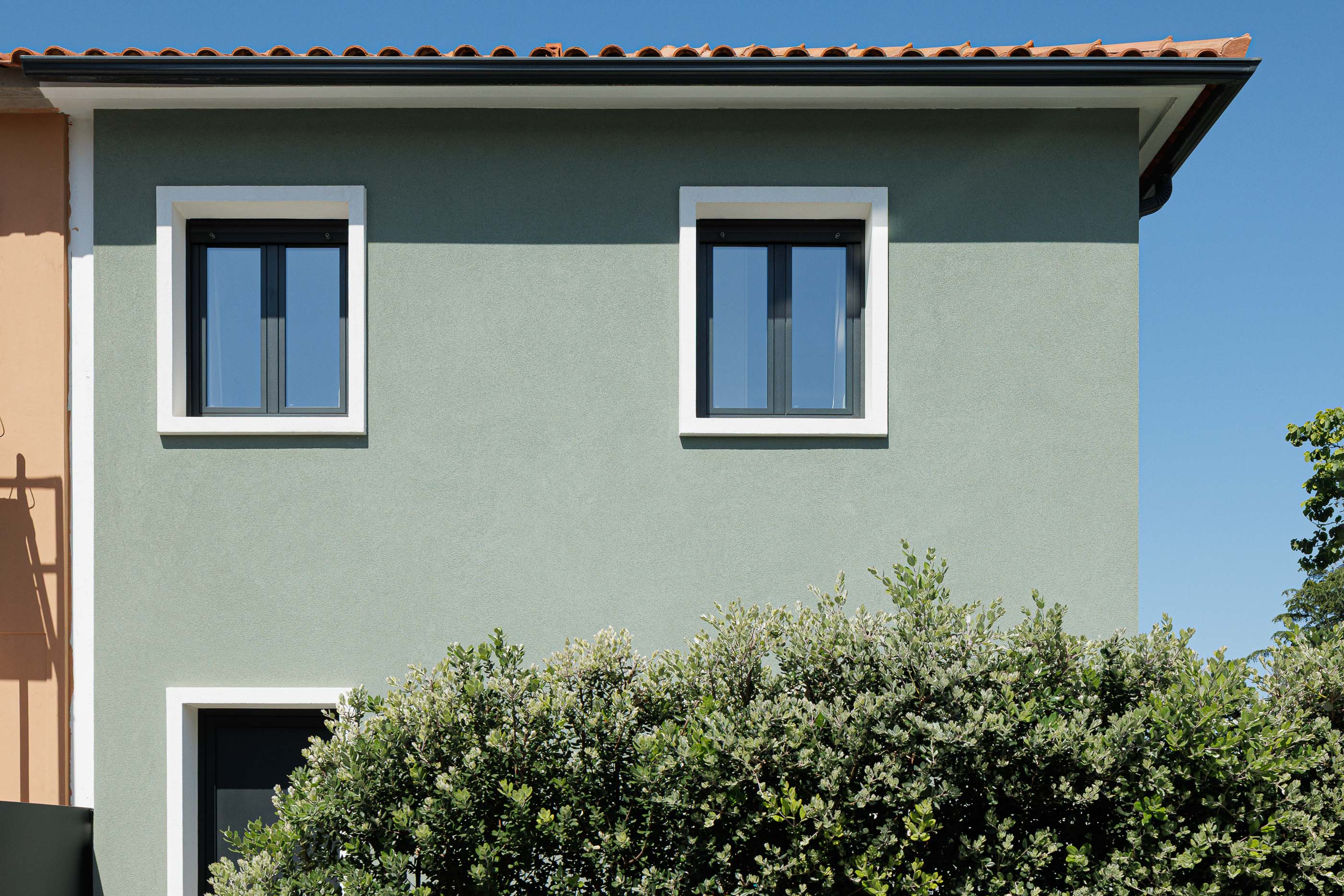 Fachada: Detalhe da fachada verde com janelas emolduradas a branco e vegetação densa. Arquitetura moderna e minimalista. — Casa Pi arquitetura ArquitectosRT em Porto