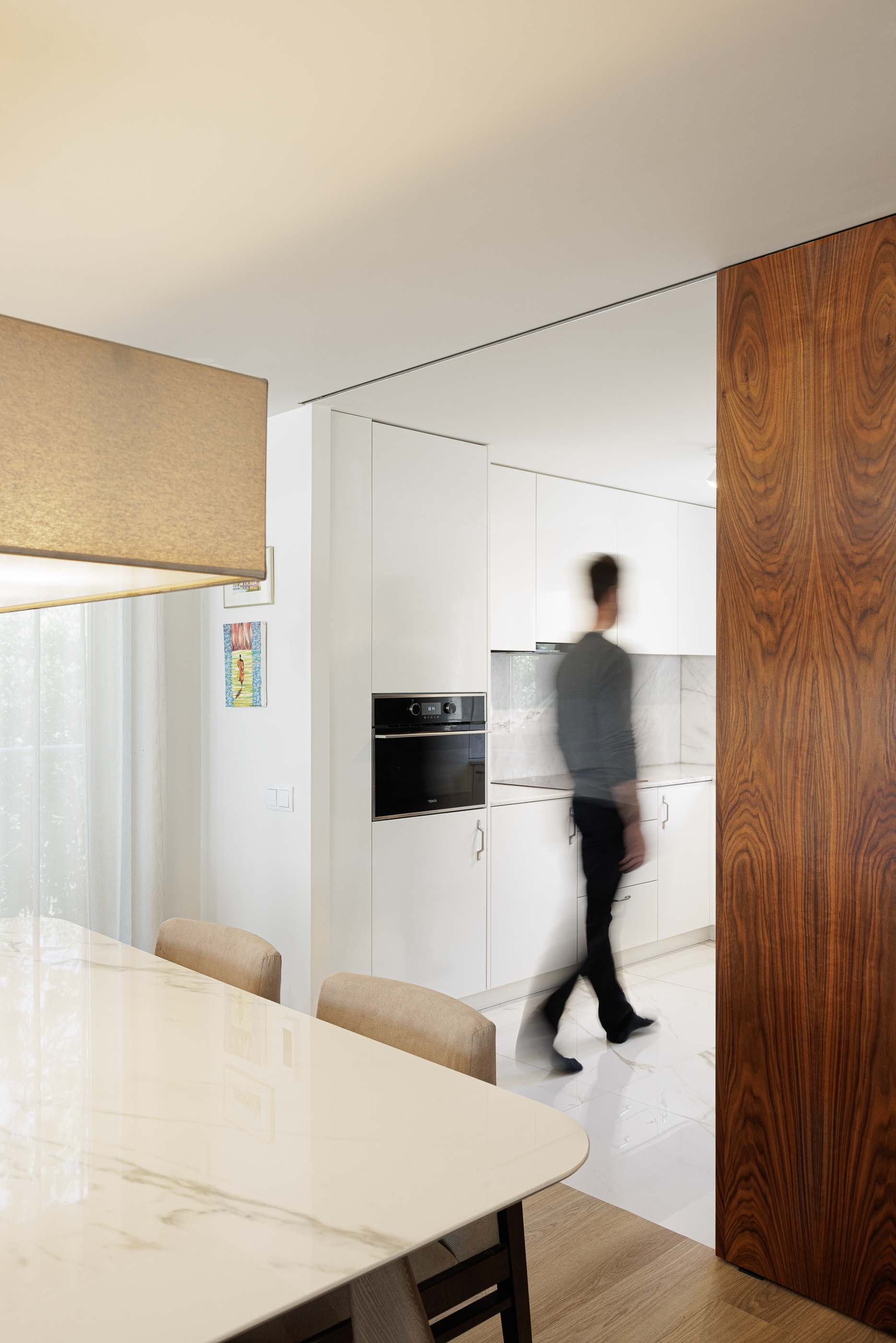 Sala de jantar/Cozinha: Vista da sala de jantar com mesa de mármore, cadeiras estofadas e cozinha branca ao fundo. — Casa Pi arquitetura ArquitectosRT em Porto