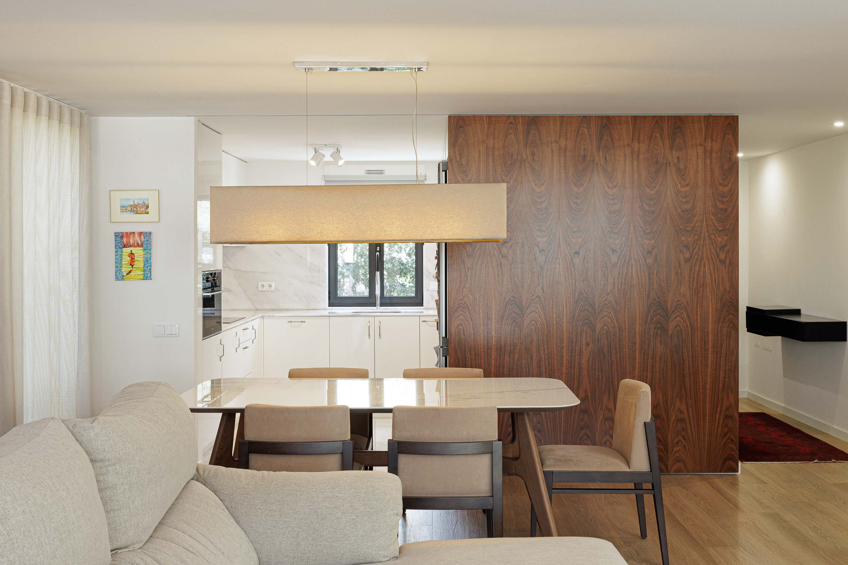 Sala de jantar/Cozinha: Sala de jantar com candeeiro retangular, parede em madeira e vista para a cozinha branca. — Casa Pi arquitetura ArquitectosRT em Porto
