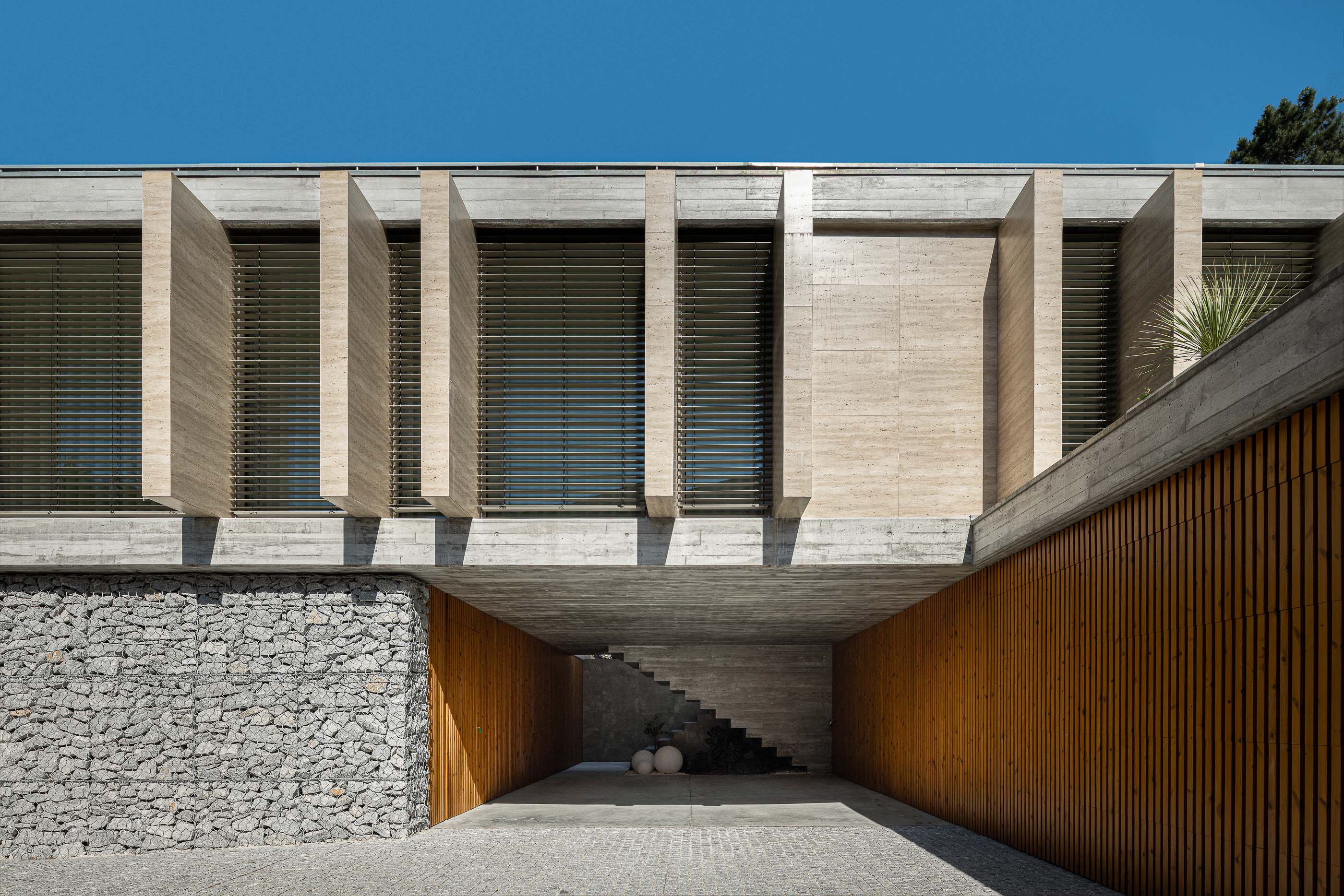 Entrada: Fachada em betão e pedra, com garagem coberta e escadas de acesso ao piso superior. — Habitação CL arquitetura Artequitectos em Guimarães