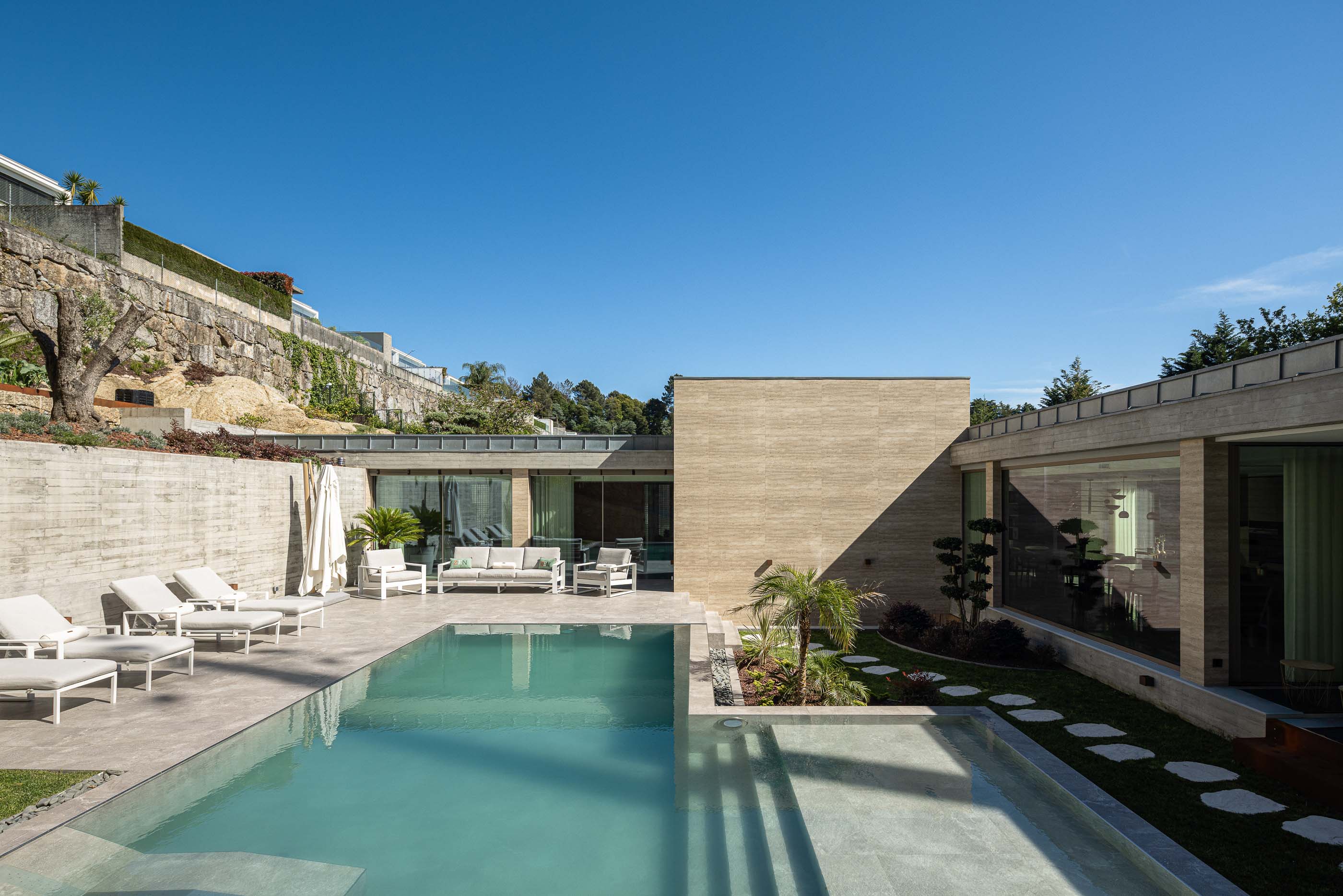 Exterior: Piscina com zona de estar, espreguiçadeiras e fachada da casa em betão. — Habitação CL arquitetura Artequitectos em Guimarães