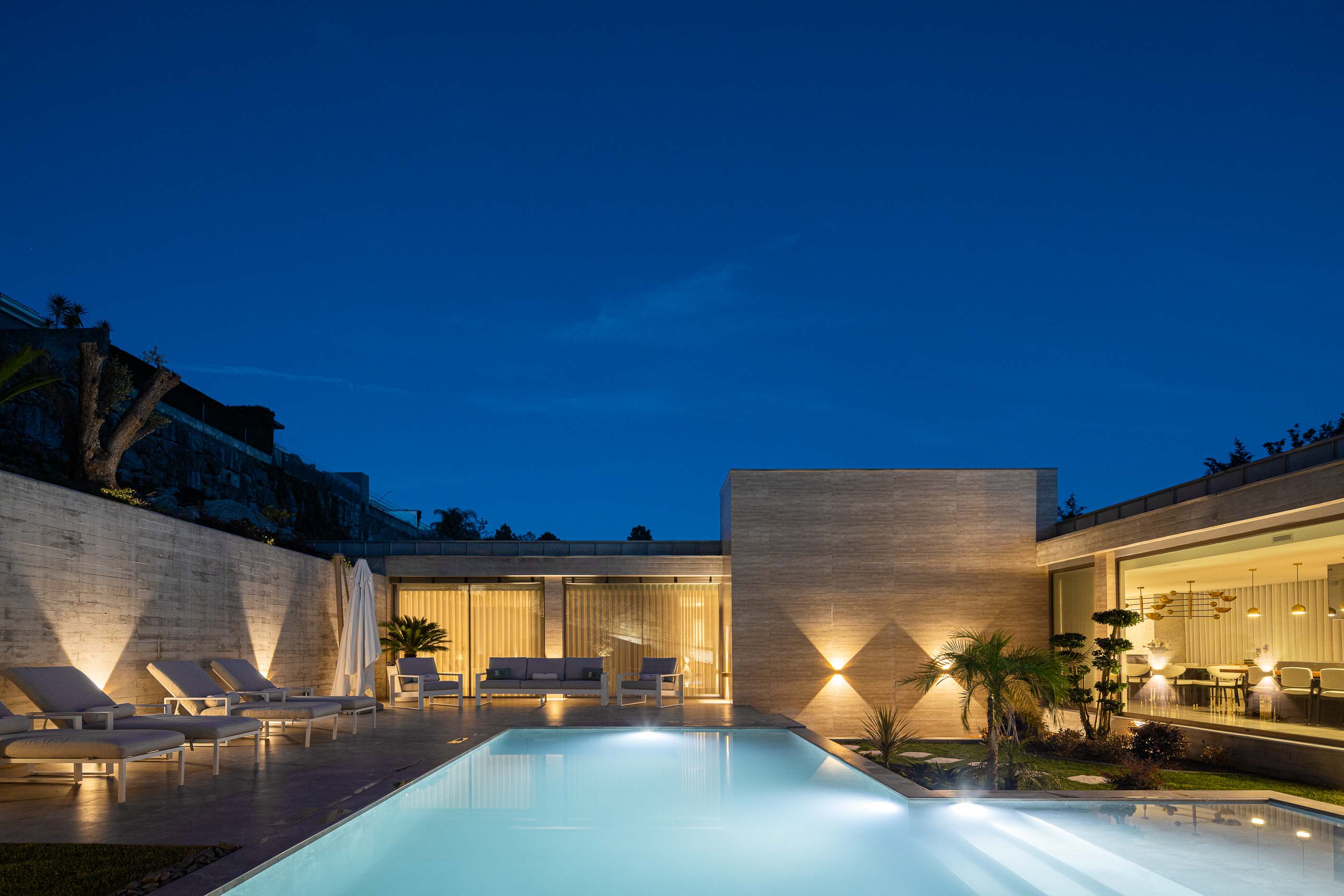 Exterior: Vista noturna da piscina com zona de estar e fachada iluminada. — Habitação CL arquitetura Artequitectos em Guimarães
