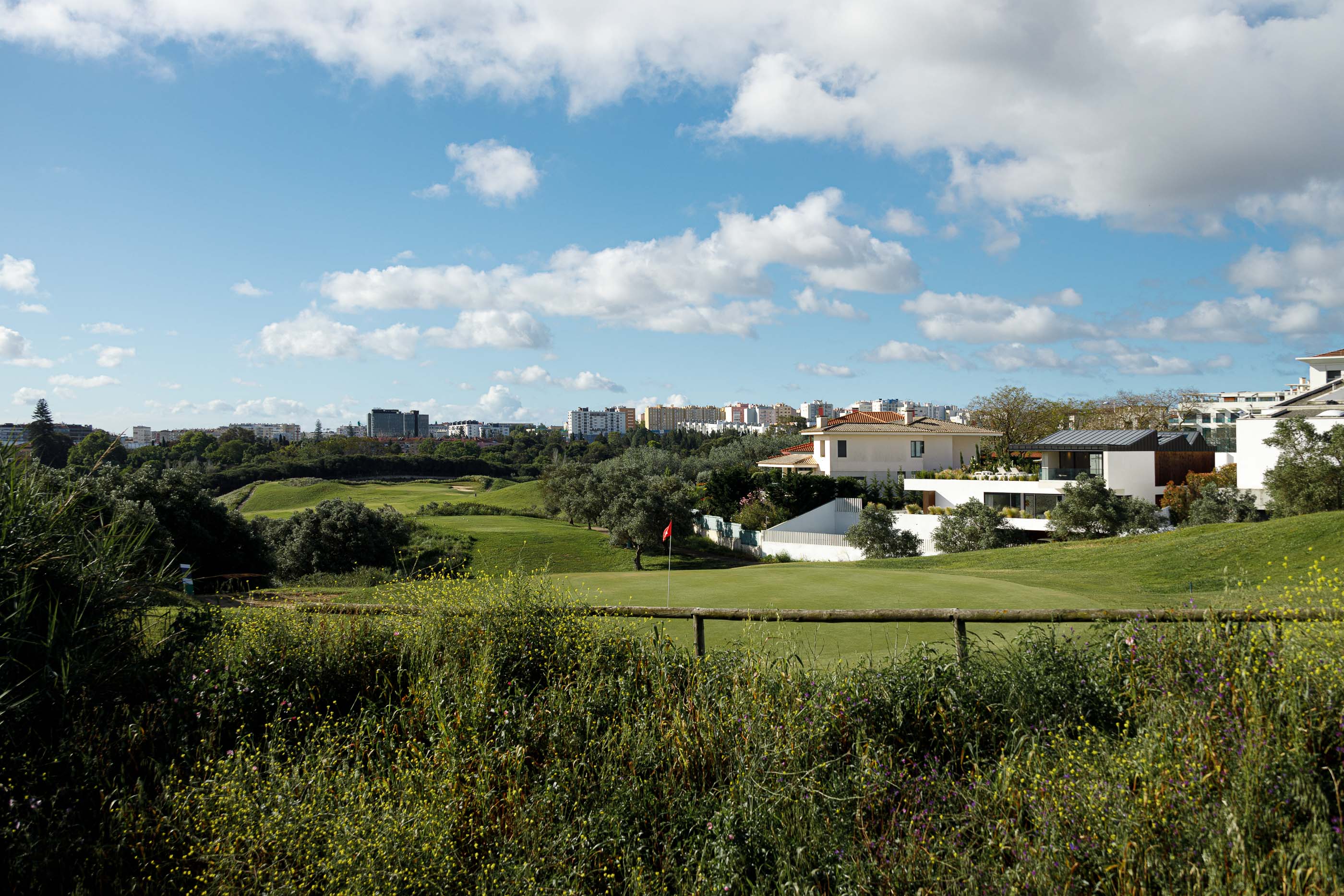 Paisagem: Vista para um campo de golfe com vegetação densa e edifícios residenciais ao fundo, sob um céu azul com nuvens. — Casa dos Alcoutins arquitetura Paulo Martins Arquitectura e Design em Lisboa