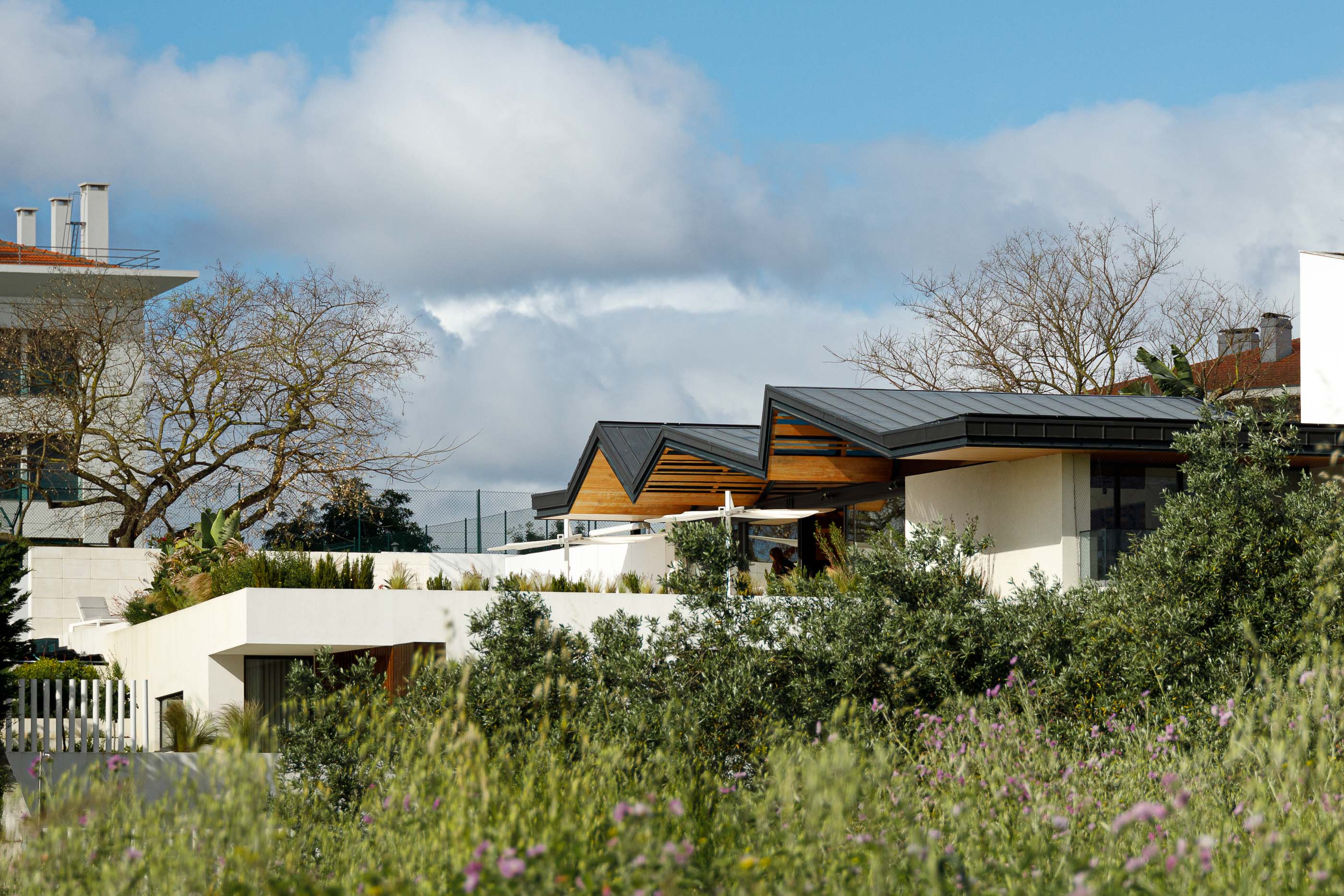 Exterior: Fachada moderna com telhados angulares, revestimento em madeira e paredes brancas, integrada na vegetação circundante. — Casa dos Alcoutins arquitetura Paulo Martins Arquitectura e Design em Lisboa