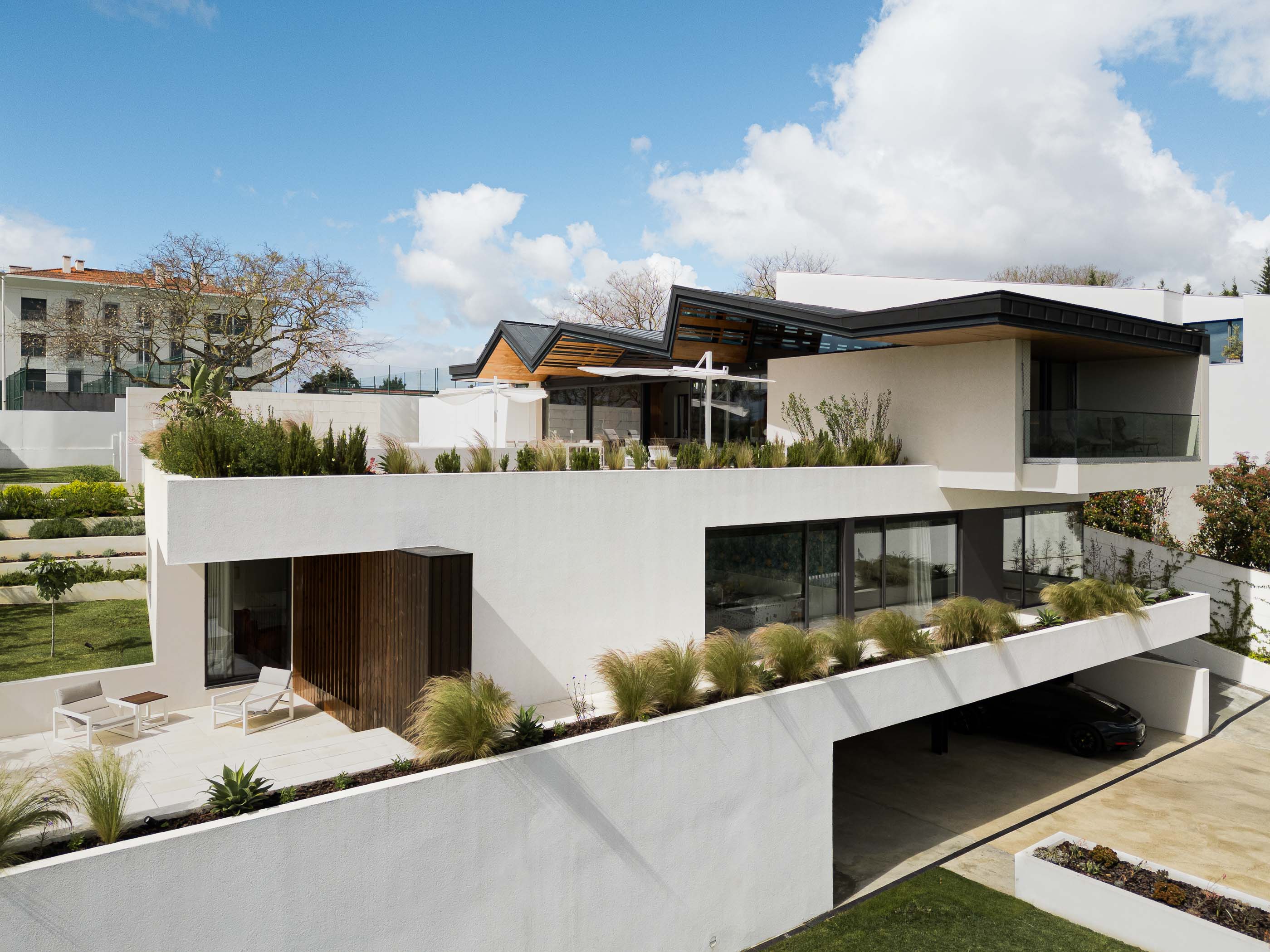 Exterior: Vista da fachada branca com detalhes em madeira, terraço com mobiliário e garagem aberta, com vegetação. — Casa dos Alcoutins arquitetura Paulo Martins Arquitectura e Design em Lisboa