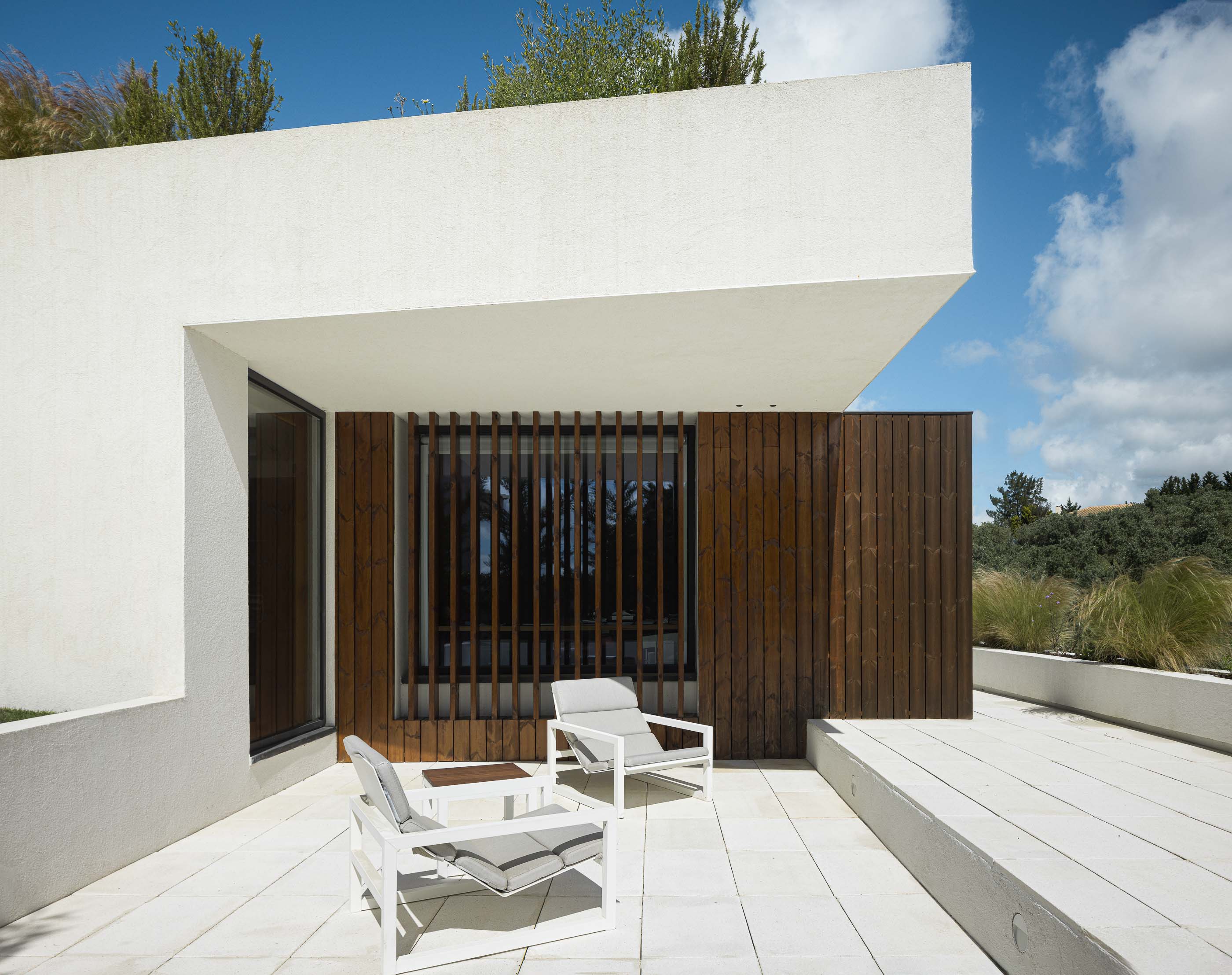 Terraço: Espaço exterior com pavimento em pedra, cadeiras de design moderno e painel de madeira vertical. — Casa dos Alcoutins arquitetura Paulo Martins Arquitectura e Design em Lisboa
