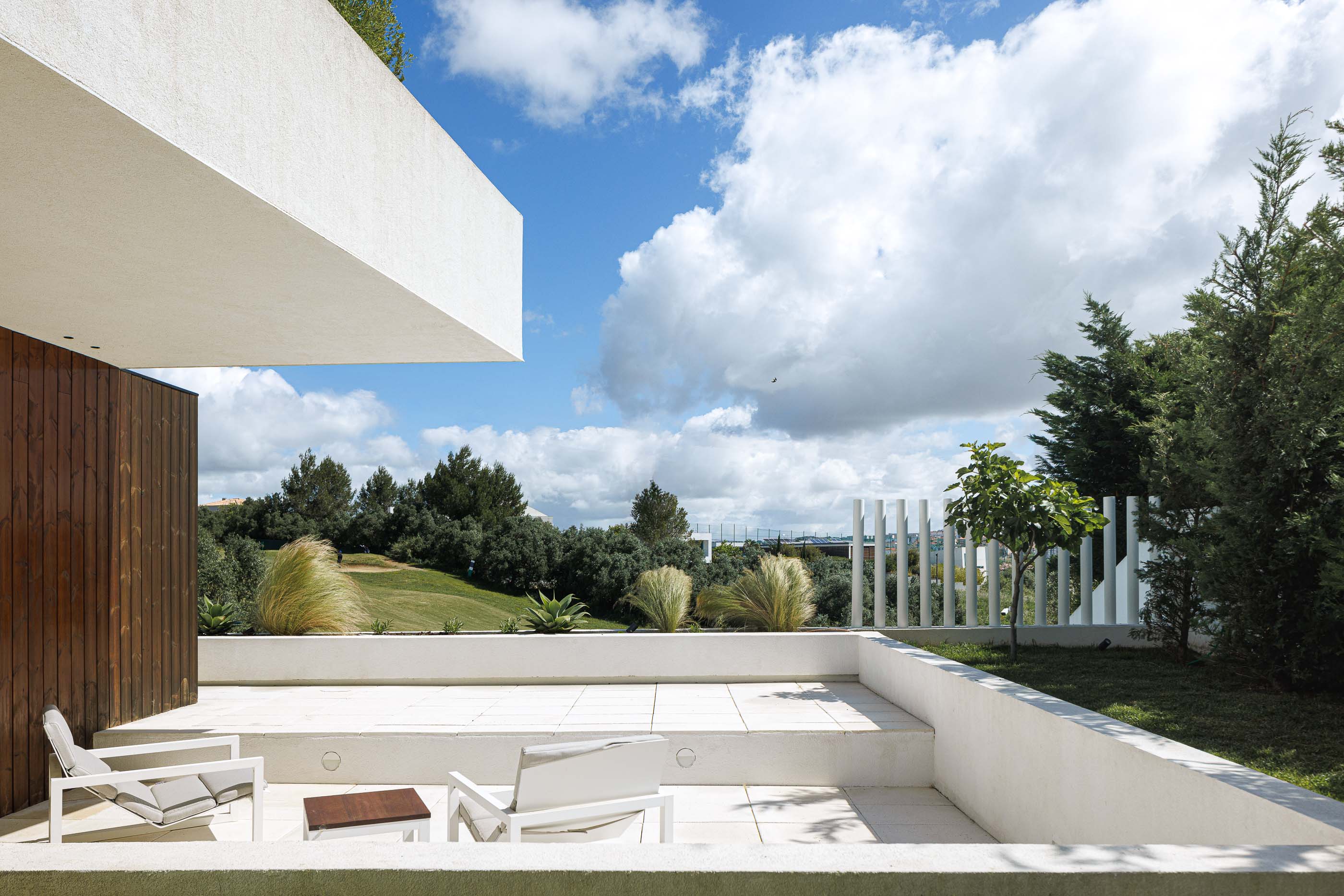 Terraço: Zona de estar exterior com cadeiras brancas, mesa de apoio e vista para o campo de golfe. — Casa dos Alcoutins arquitetura Paulo Martins Arquitectura e Design em Lisboa