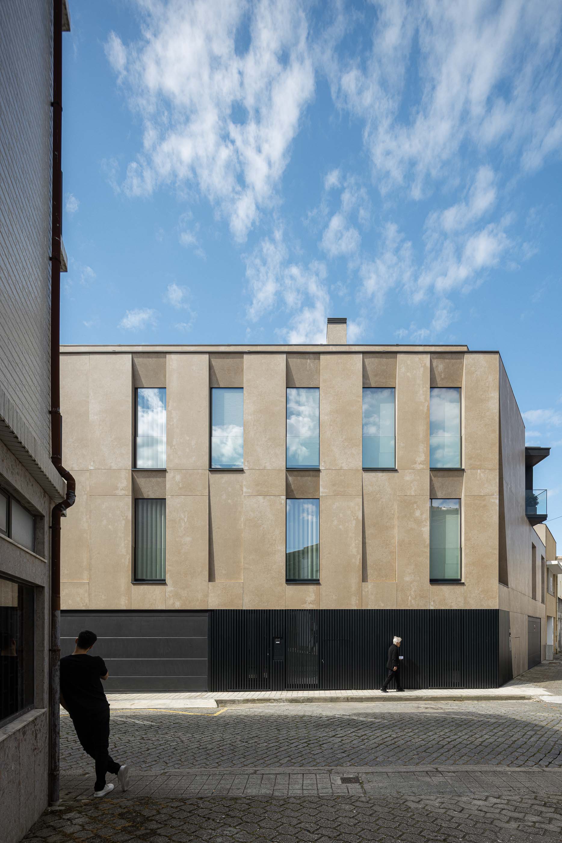 Fachada: Vista frontal do edifício com fachada em tons terra, janelas verticais e portões pretos. — Reabilitação Poveira - Edifício FS11 arquitetura Urbanpolis em Póvoa de Varzim