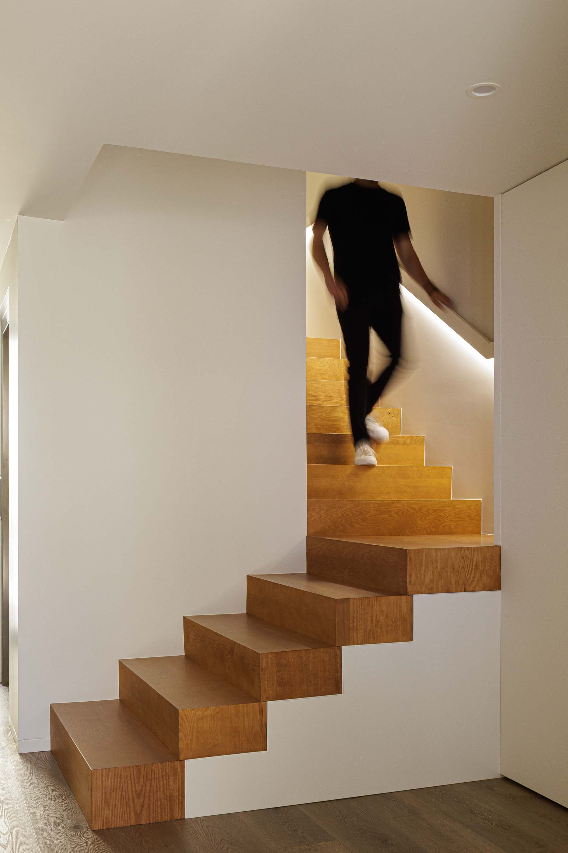 Escada: Escada interior em madeira clara com iluminação embutida na parede branca. — Reabilitação Poveira - Edifício FS11 arquitetura Urbanpolis em Póvoa de Varzim