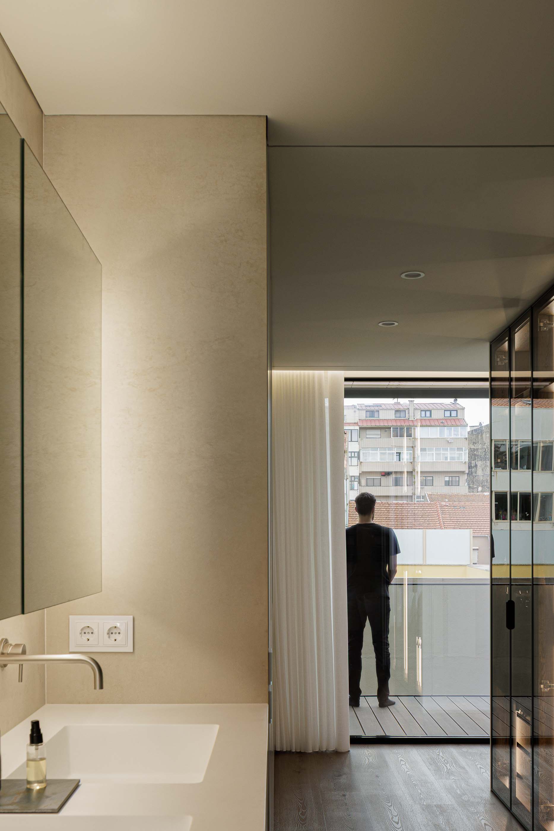 Casa de banho: Lavatório branco com espelho iluminado e vista para a varanda envidraçada. — Reabilitação Poveira - Edifício FS11 arquitetura Urbanpolis em Póvoa de Varzim