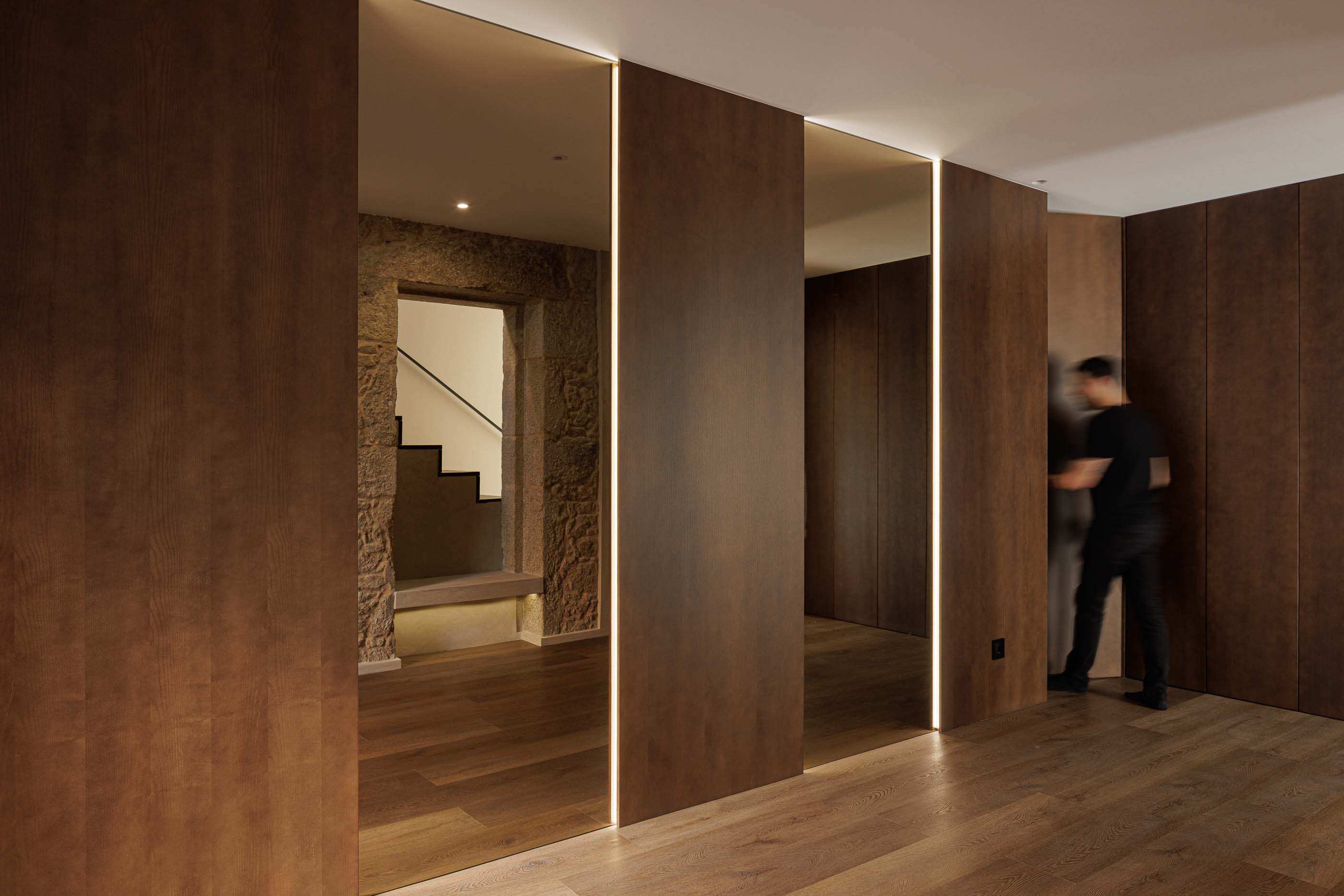 Hall de entrada: Espaço com paredes em madeira, espelhos com iluminação e detalhe da parede em pedra original. — Reabilitação Poveira - Edifício FS11 arquitetura Urbanpolis em Póvoa de Varzim