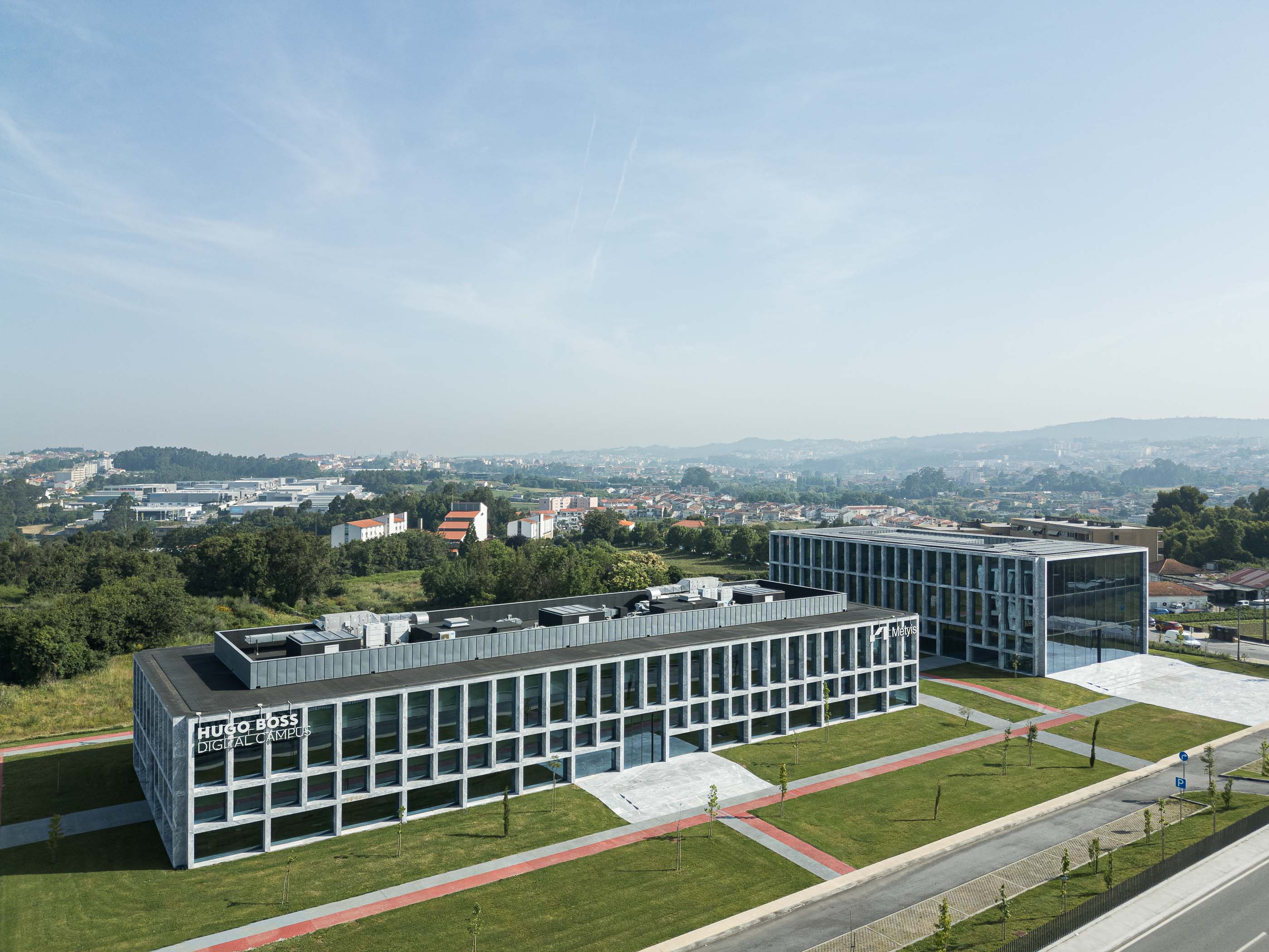 Exterior: Vista aérea do Metyis Digital Campus, com os dois edifícios, relvado e a cidade ao fundo. — Metyis Digital Campus arquitetura UNUM - Oficina de Arquitetura em Gondomar