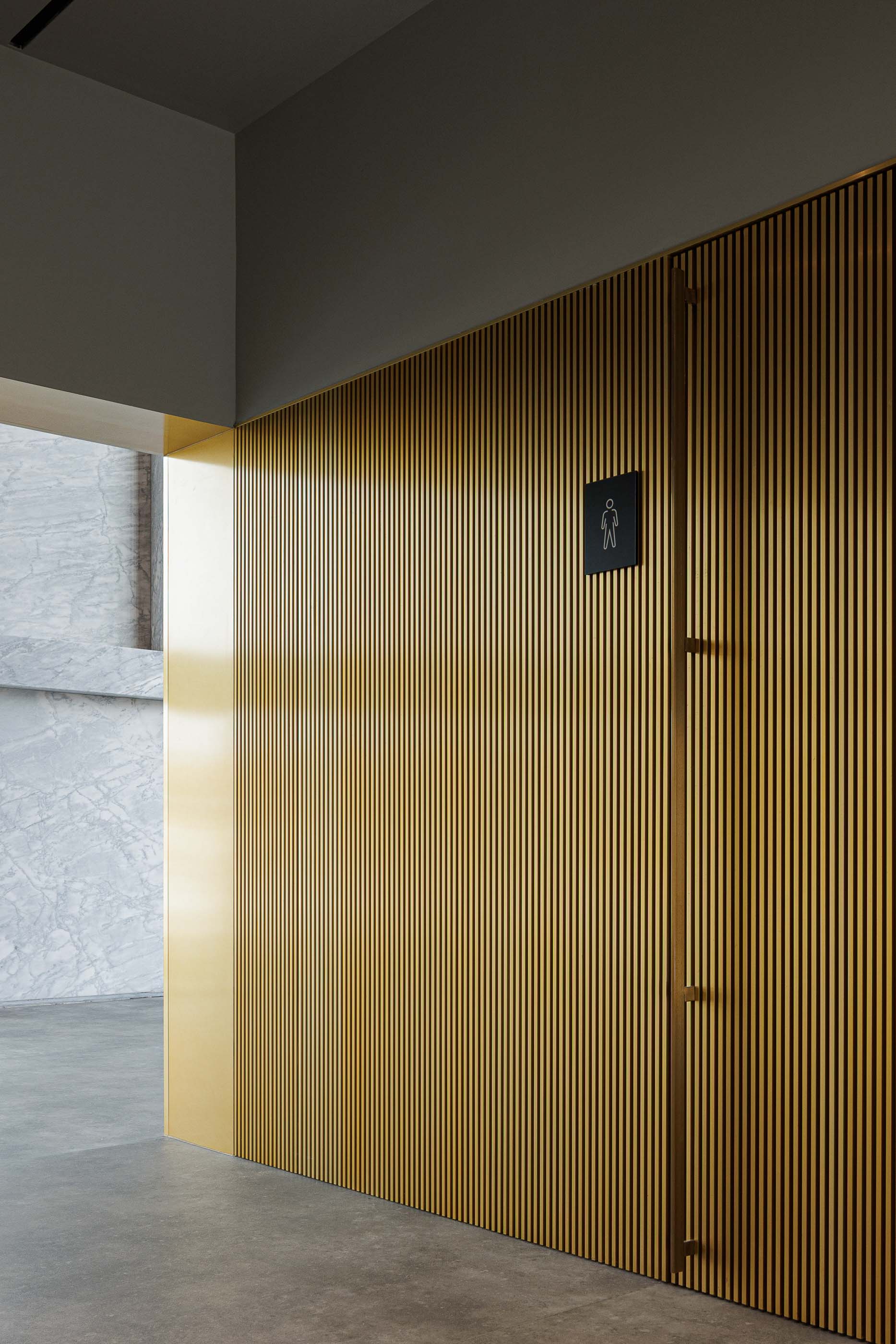 Casa de banho: Detalhe da porta da casa de banho masculina com revestimento em madeira ripada e detalhes em dourado. — Metyis Digital Campus arquitetura UNUM - Oficina de Arquitetura em Gondomar