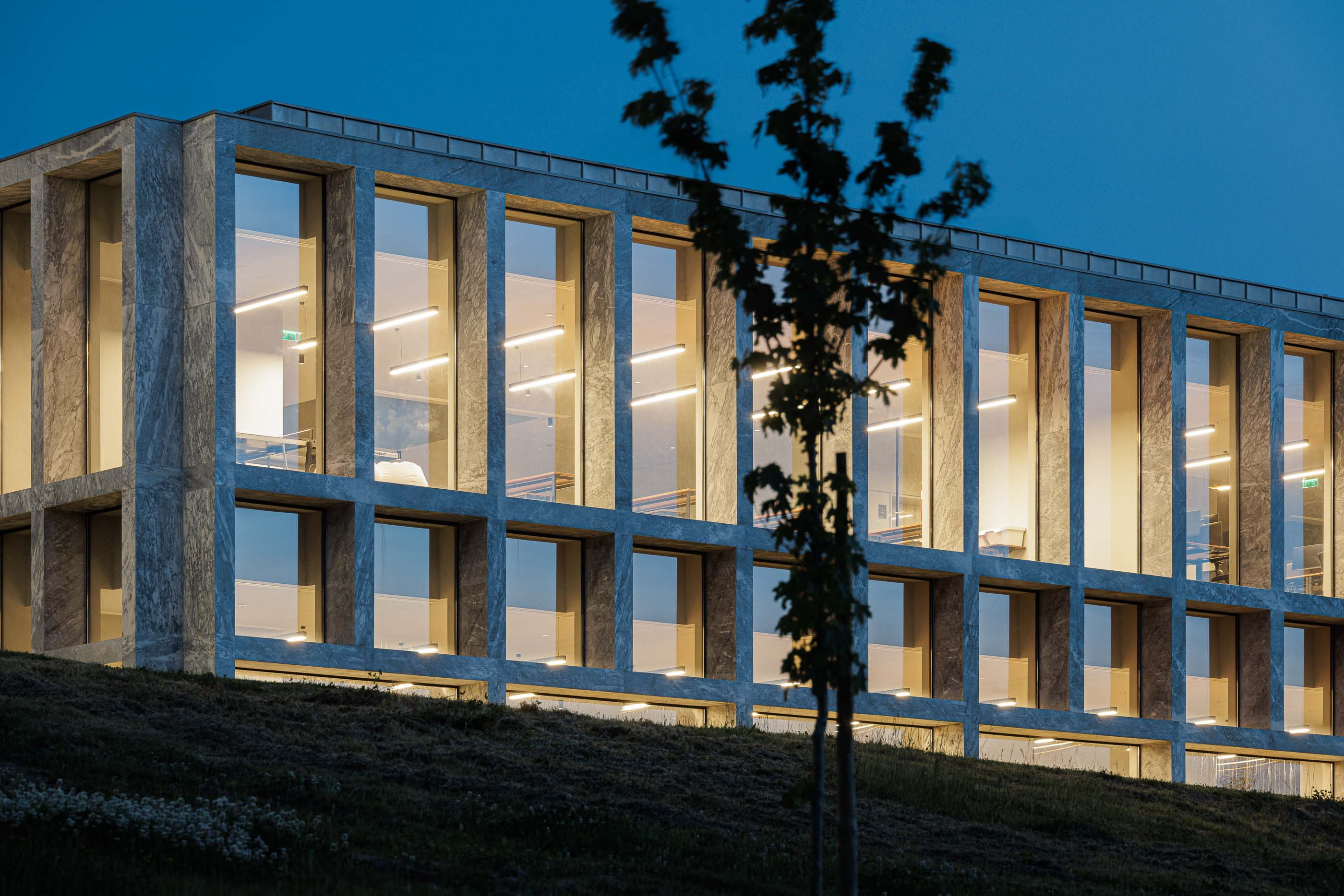 Edifício: Detalhe noturno da fachada em mármore com janelas iluminadas. — Metyis Digital Campus arquitetura UNUM - Oficina de Arquitetura em Gondomar