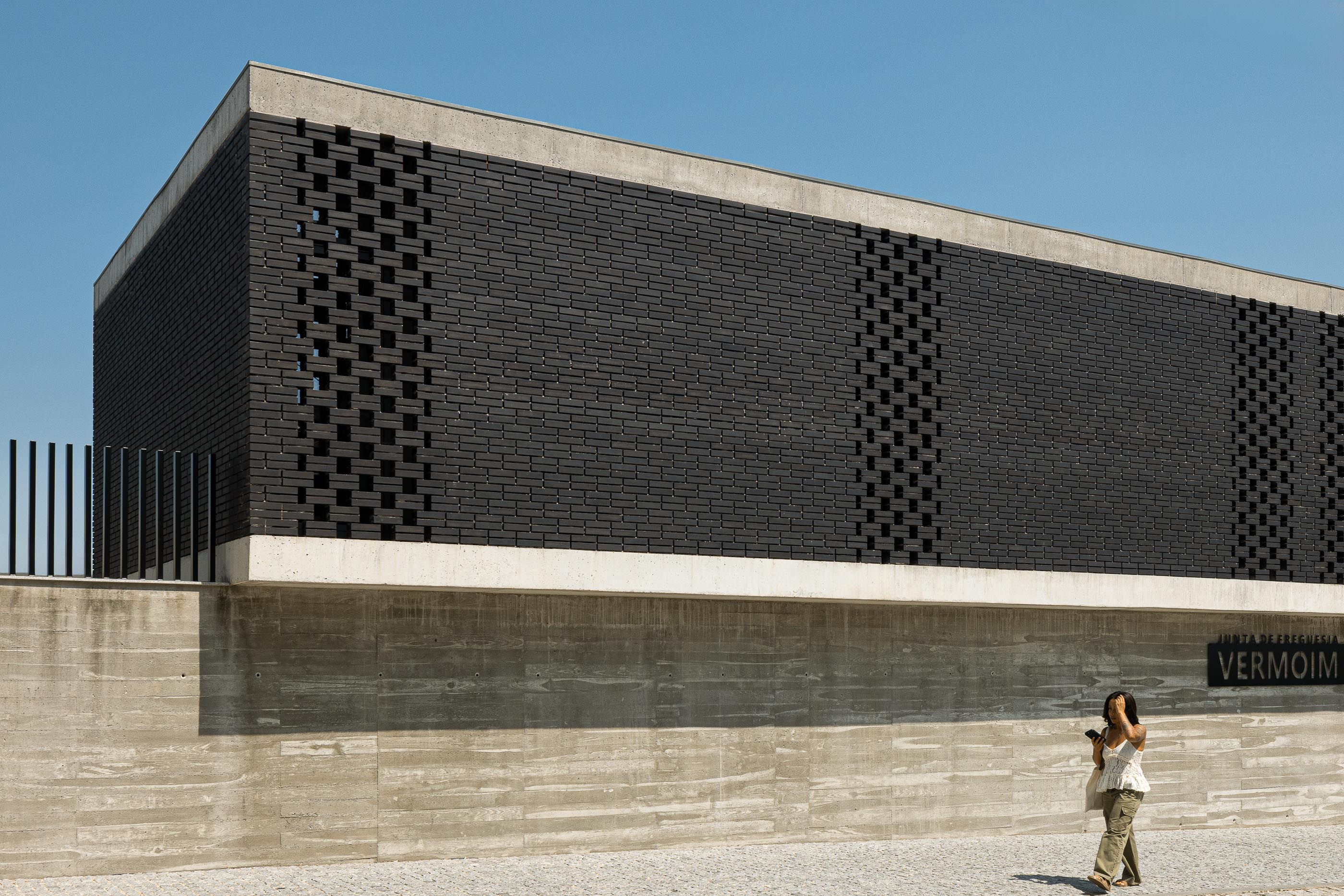 Fachada: Edifício moderno com tijolo preto perfurado e base em betão aparente, sob céu azul. Pessoa a caminhar em primeiro plano. — Junta de Freguesia de Vermoim arquitetura Rui Mendes Ribeiro Arquitecto em Vila Nova de Famalicão