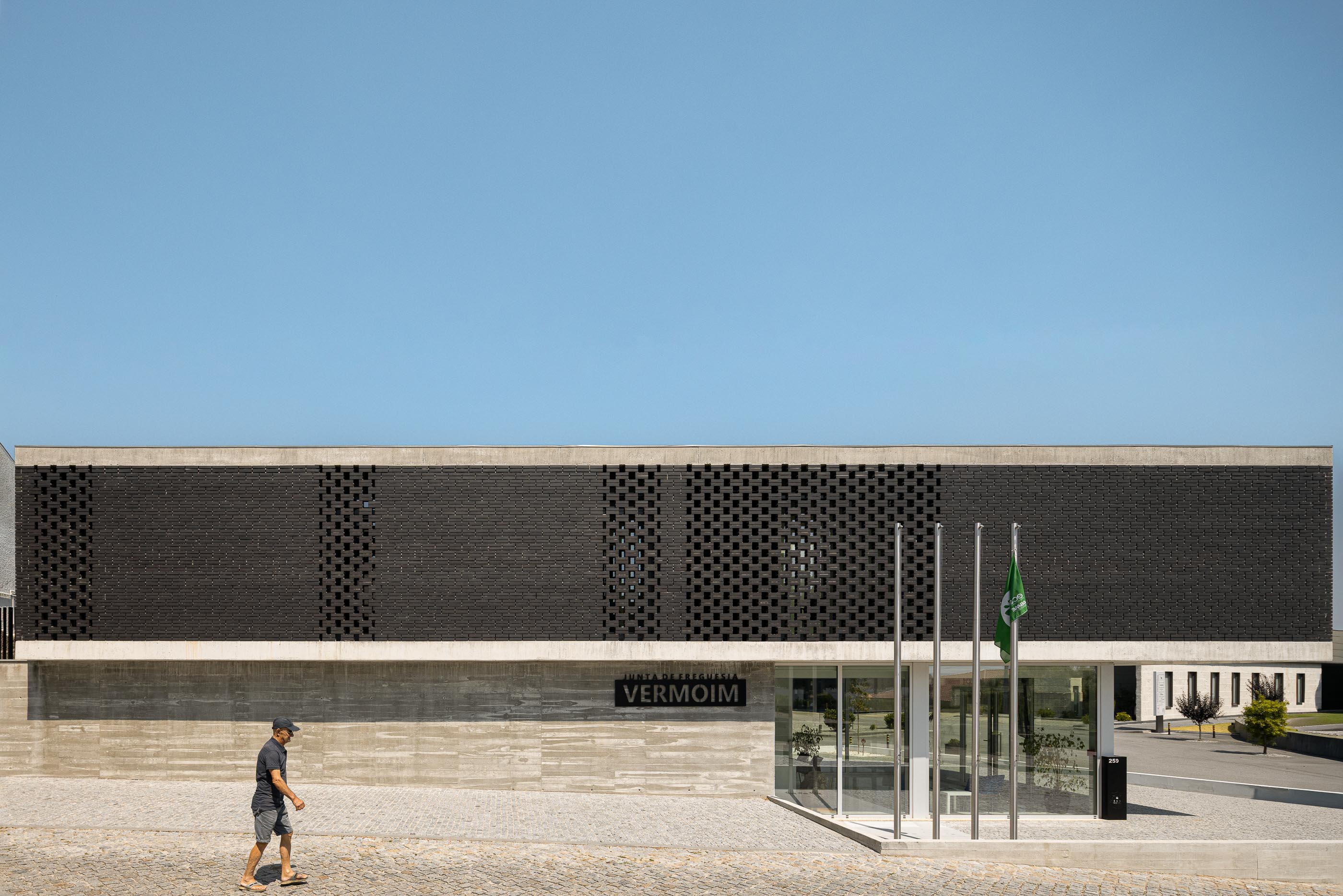 Fachada: Edifício da Junta de Freguesia com tijolo preto, base em betão e bandeiras. Calçada portuguesa em frente. — Junta de Freguesia de Vermoim arquitetura Rui Mendes Ribeiro Arquitecto em Vila Nova de Famalicão