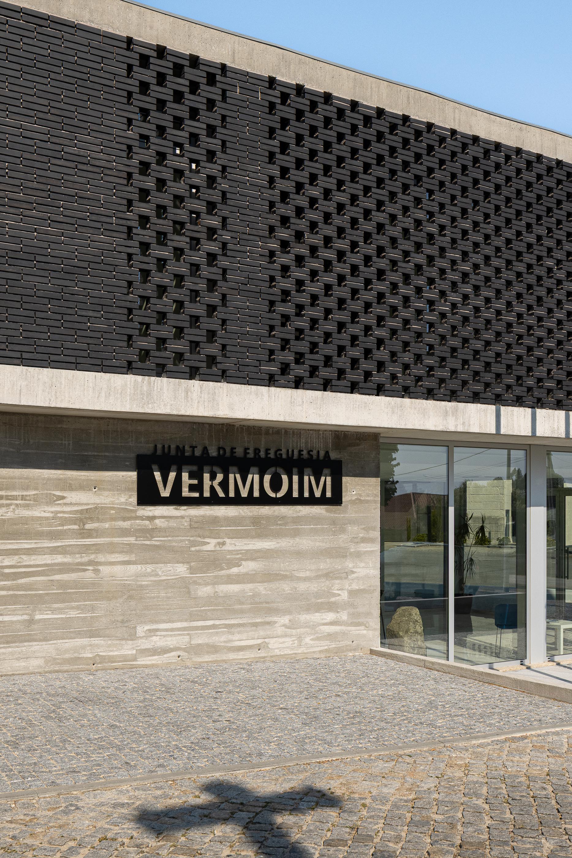 Fachada: Detalhe da fachada da Junta de Freguesia, com tijolo preto, betão aparente e placa identificativa. — Junta de Freguesia de Vermoim arquitetura Rui Mendes Ribeiro Arquitecto em Vila Nova de Famalicão