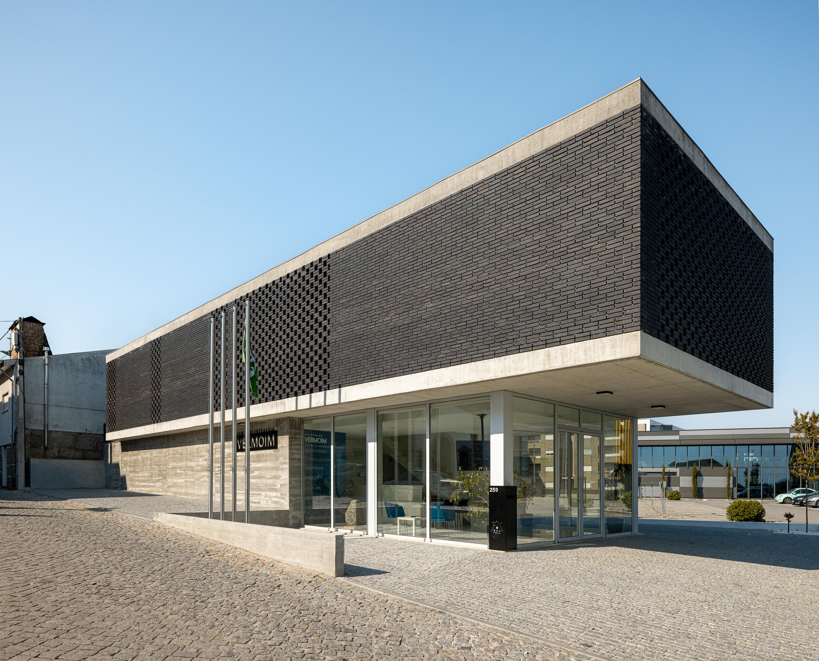 Fachada: Vista angular do edifício da Junta de Freguesia, com fachada envidraçada e tijolo preto. — Junta de Freguesia de Vermoim arquitetura Rui Mendes Ribeiro Arquitecto em Vila Nova de Famalicão