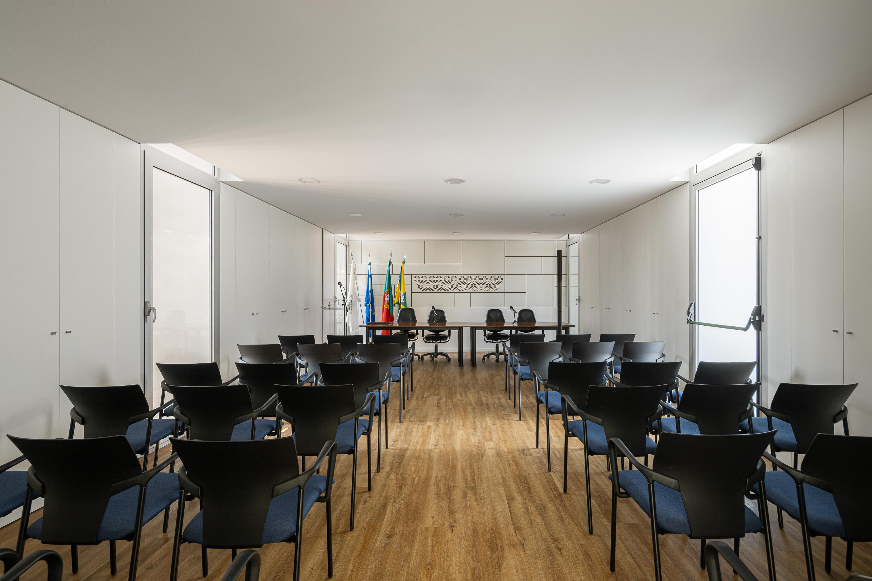 Sala de Reuniões: Cadeiras azuis dispostas em frente a mesa de madeira, sob luz branca. — Junta de Freguesia de Vermoim arquitetura Rui Mendes Ribeiro Arquitecto em Vila Nova de Famalicão