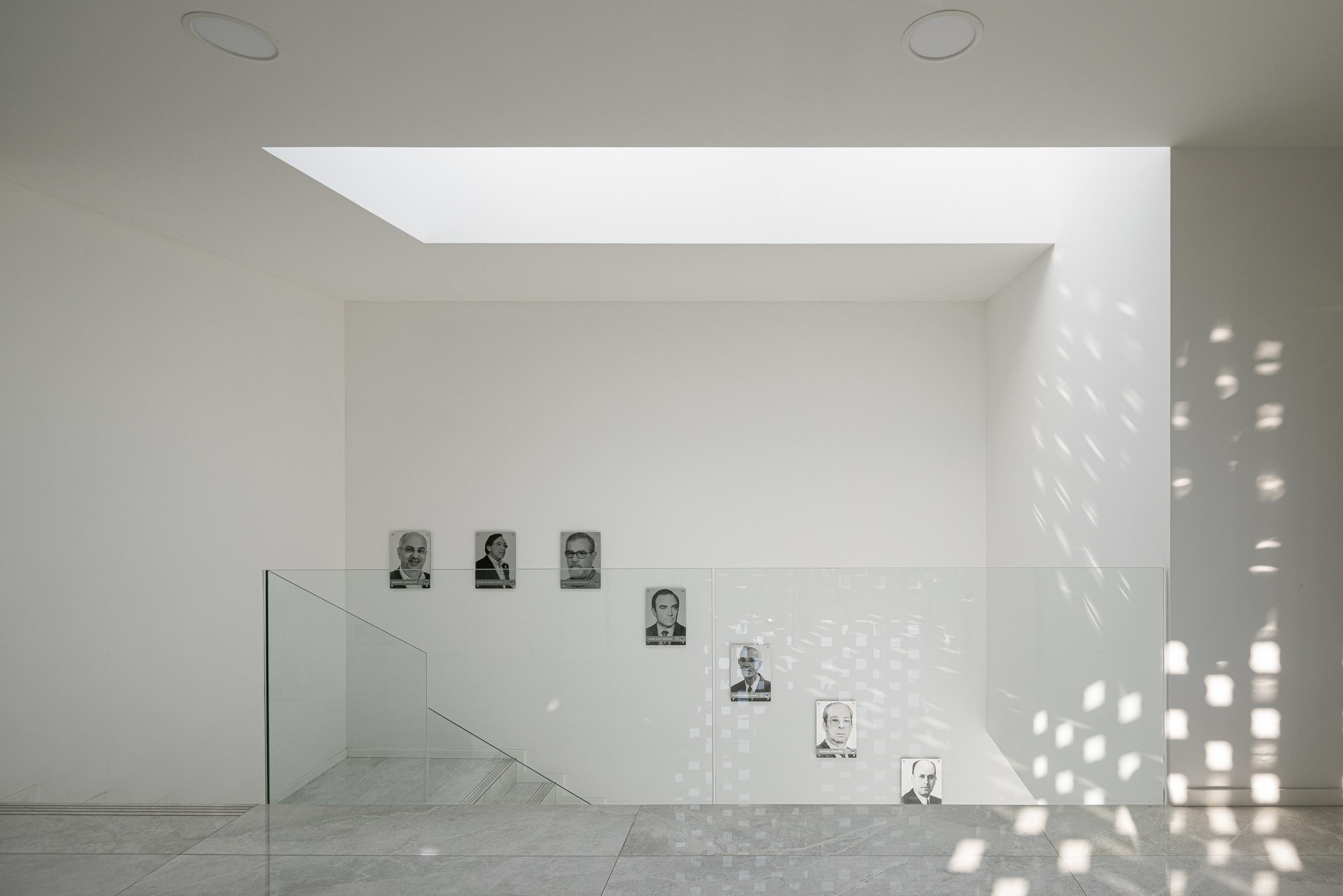 Escadaria: Escadas com corrimão de vidro, paredes brancas e claraboia, com retratos na parede. — Junta de Freguesia de Vermoim arquitetura Rui Mendes Ribeiro Arquitecto em Vila Nova de Famalicão