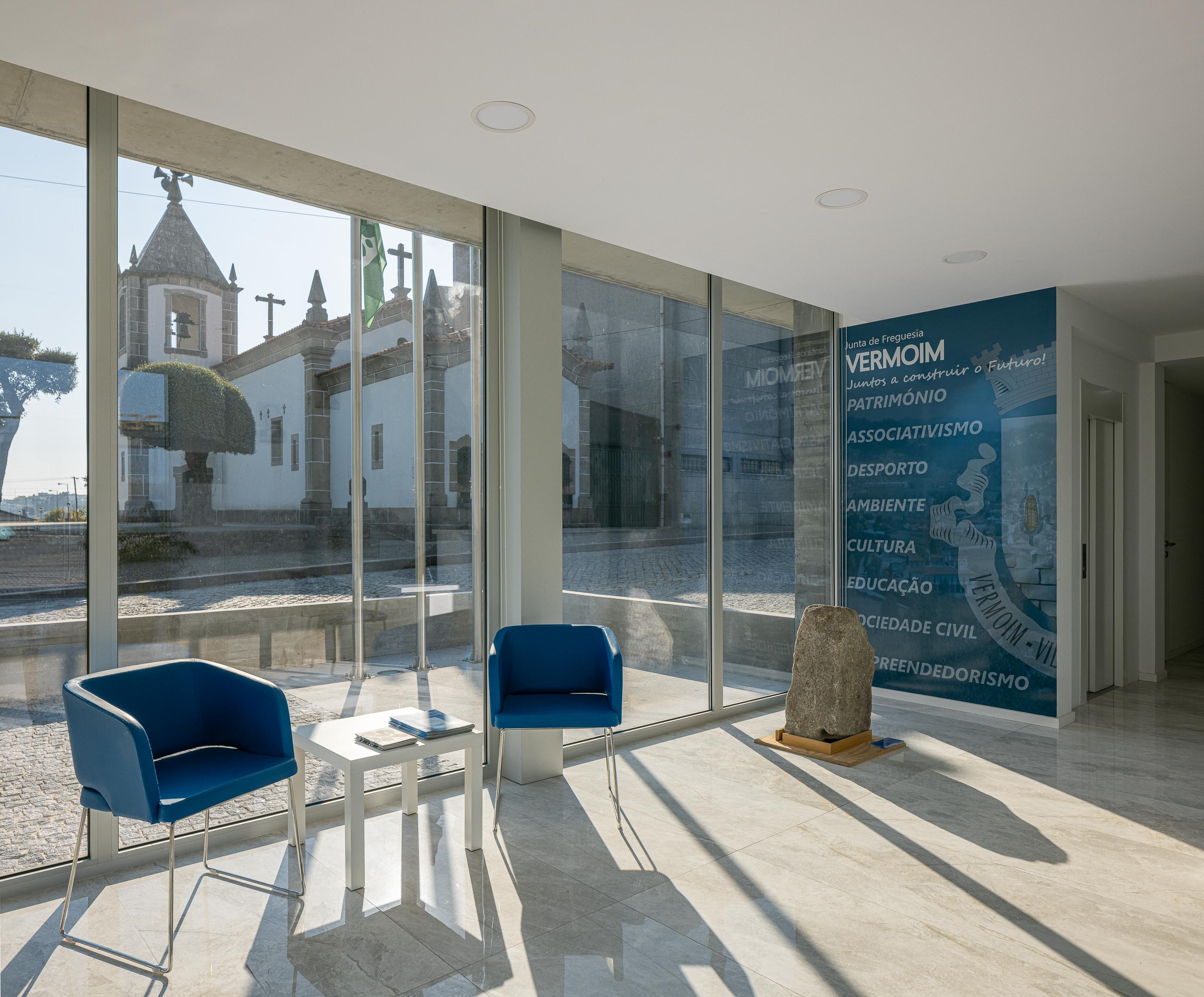 Sala de Espera: Cadeiras azuis numa sala de espera luminosa, com vista para a igreja e pavimento em mármore. — Junta de Freguesia de Vermoim arquitetura Rui Mendes Ribeiro Arquitecto em Vila Nova de Famalicão