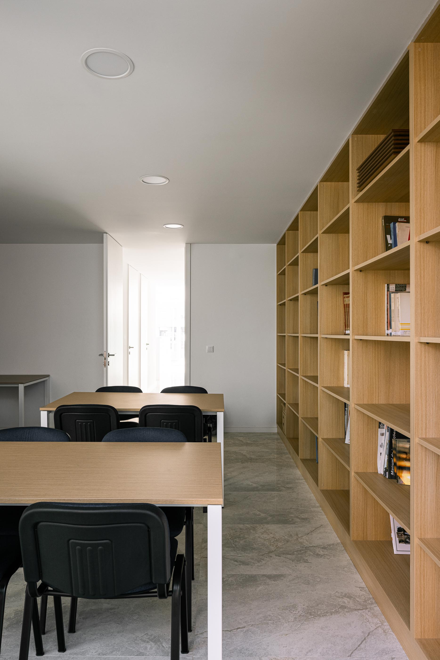 Biblioteca: Estantes de madeira claras cheias de livros, mesas de estudo e cadeiras pretas, com pavimento cinza. — Junta de Freguesia de Vermoim arquitetura Rui Mendes Ribeiro Arquitecto em Vila Nova de Famalicão