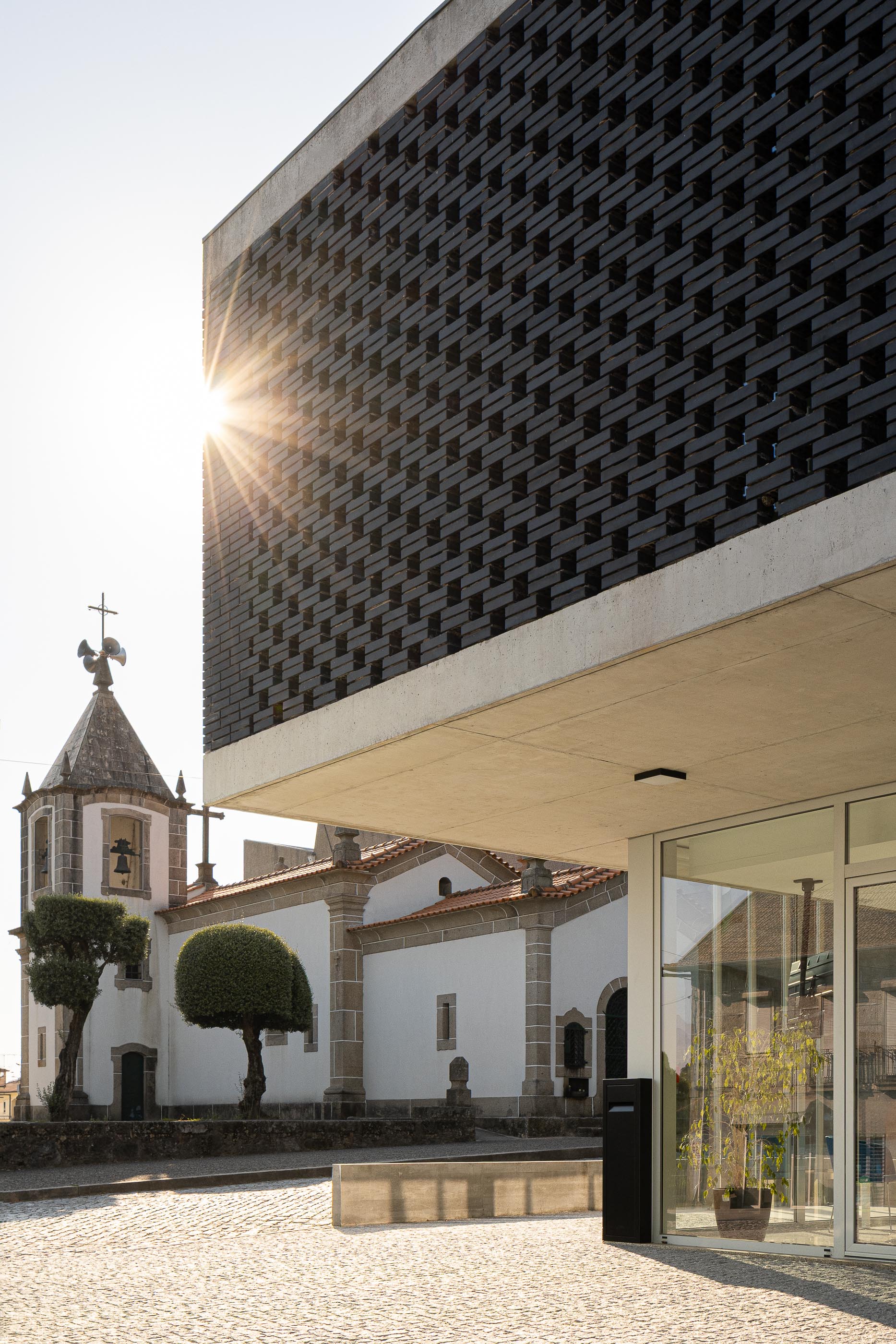 Exterior: Edifício da Junta de Freguesia com tijolo preto, ao lado da igreja matriz, com luz solar intensa. — Junta de Freguesia de Vermoim arquitetura Rui Mendes Ribeiro Arquitecto em Vila Nova de Famalicão