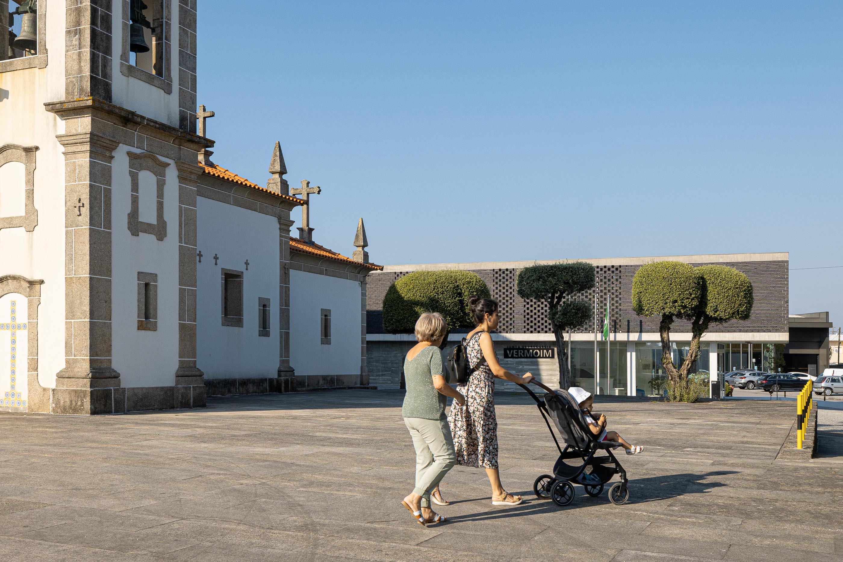 Exterior: Igreja matriz e edifício da Junta de Freguesia vistos da praça, com pessoas a passear. — Junta de Freguesia de Vermoim arquitetura Rui Mendes Ribeiro Arquitecto em Vila Nova de Famalicão