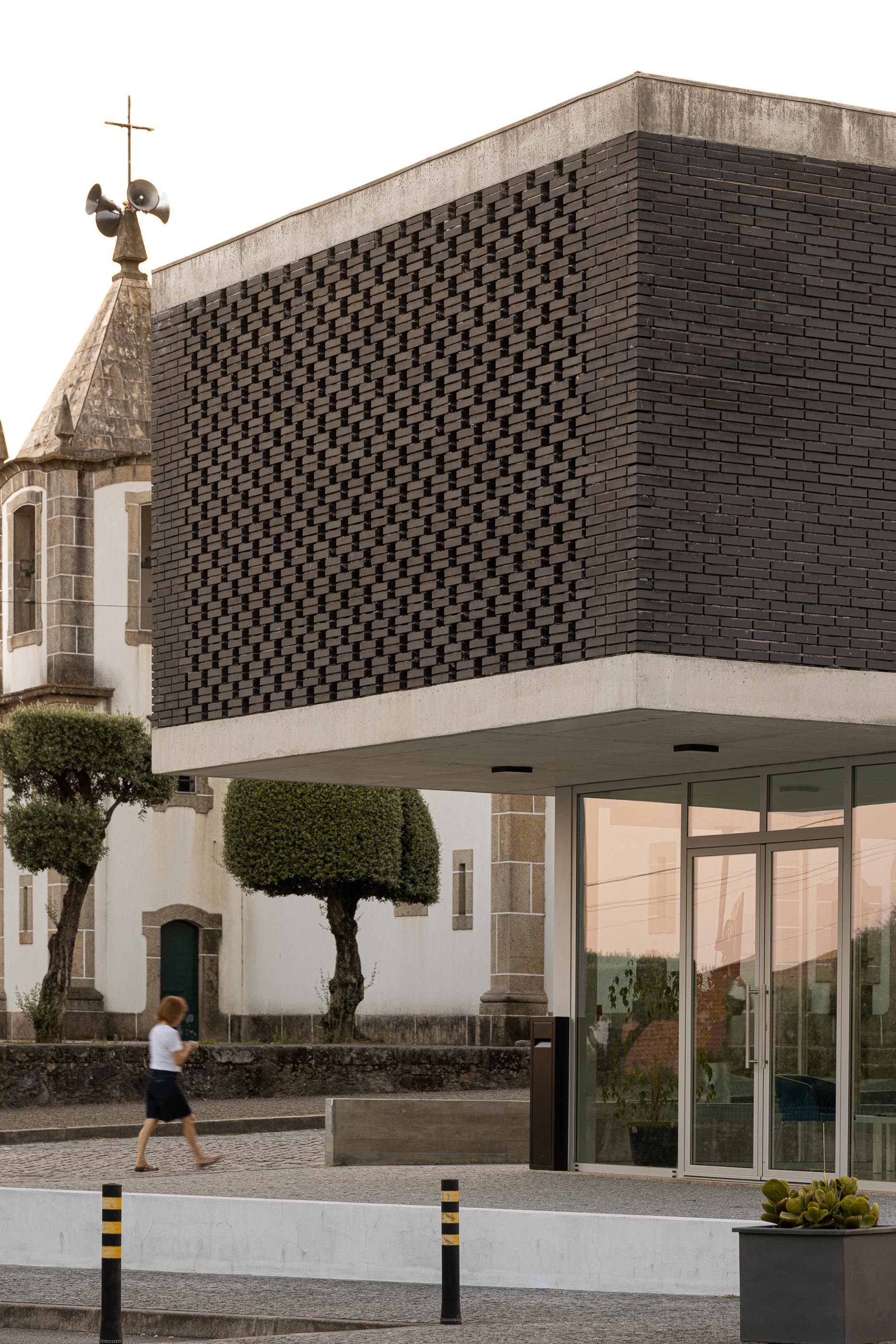 Exterior: Detalhe do edifício da Junta de Freguesia com tijolo preto e igreja ao fundo, com vegetação. — Junta de Freguesia de Vermoim arquitetura Rui Mendes Ribeiro Arquitecto em Vila Nova de Famalicão