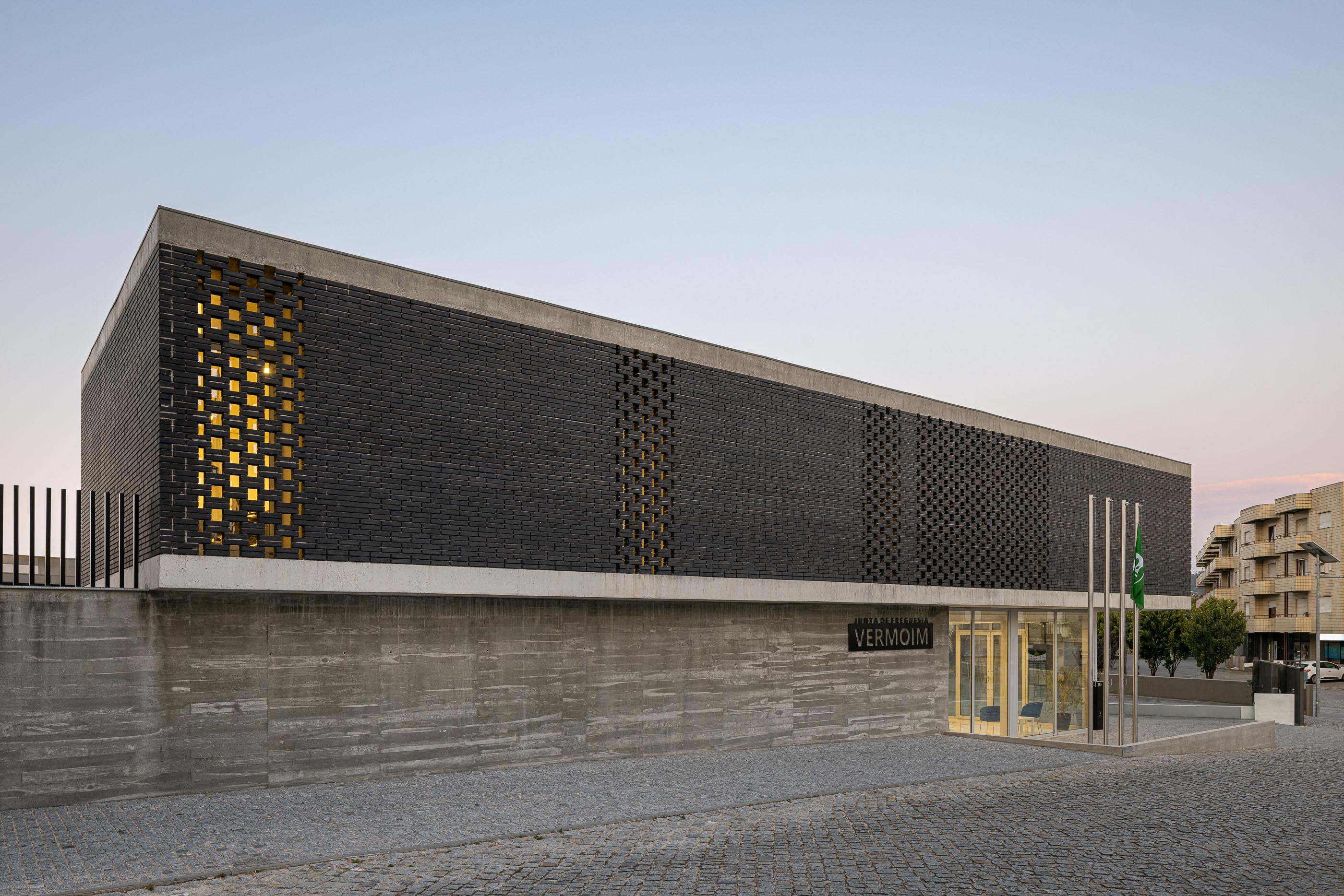 Fachada: Edifício da Junta de Freguesia com tijolo preto e base em betão, com iluminação interior. — Junta de Freguesia de Vermoim arquitetura Rui Mendes Ribeiro Arquitecto em Vila Nova de Famalicão