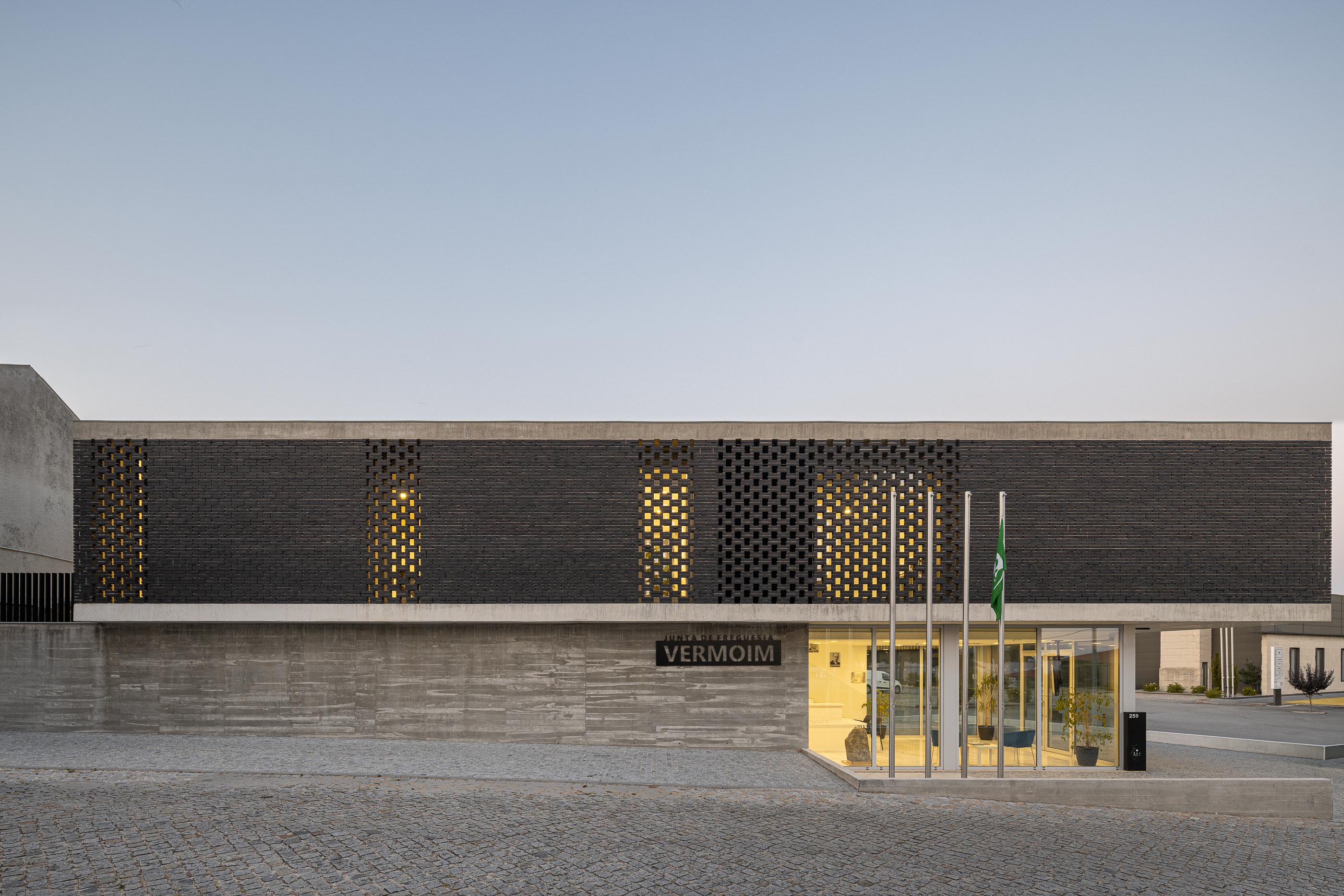 Fachada: Edifício da Junta de Freguesia com tijolo preto e base em betão, com bandeiras. — Junta de Freguesia de Vermoim arquitetura Rui Mendes Ribeiro Arquitecto em Vila Nova de Famalicão