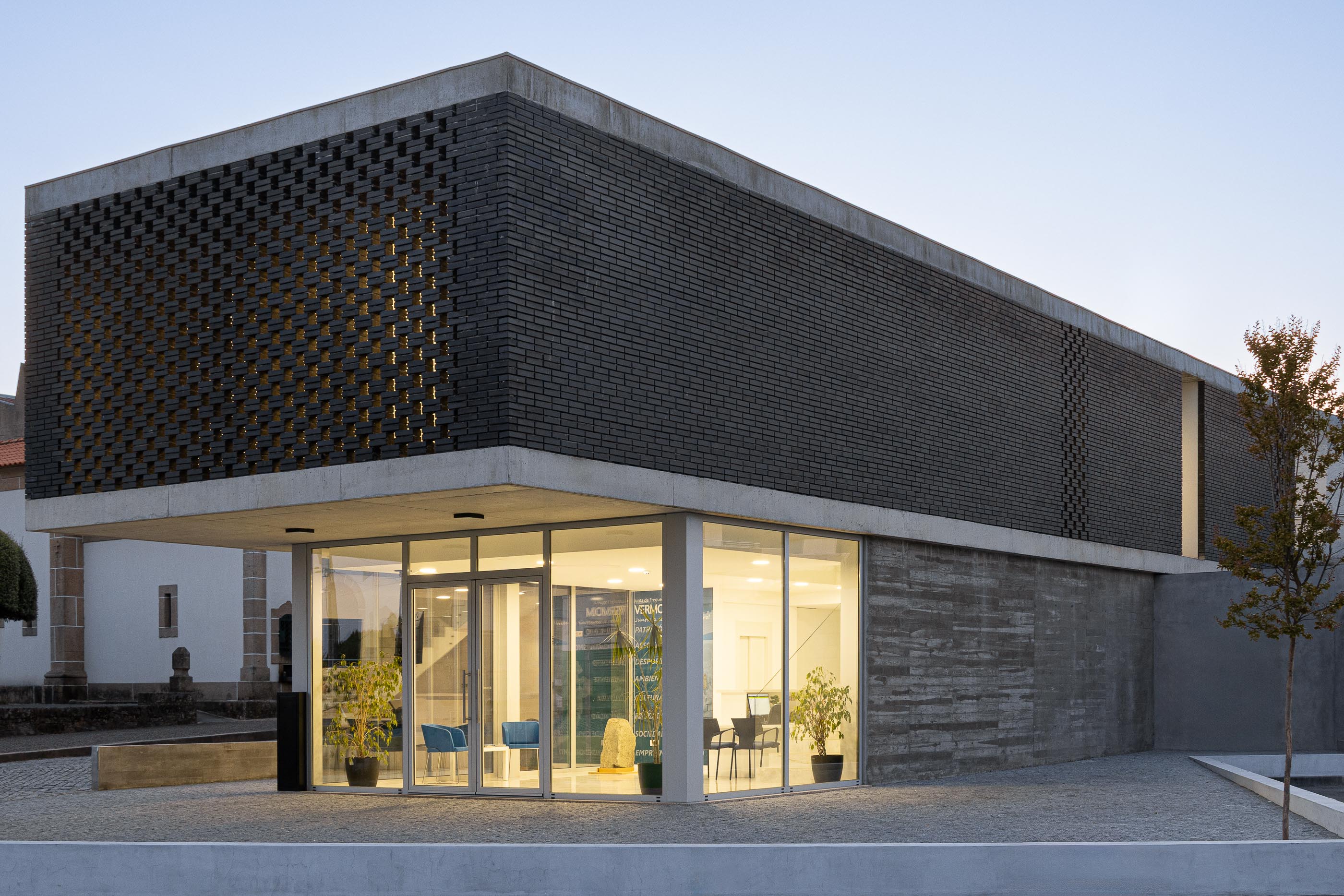 Fachada: Edifício da Junta de Freguesia com tijolo preto e fachada envidraçada, com vegetação. — Junta de Freguesia de Vermoim arquitetura Rui Mendes Ribeiro Arquitecto em Vila Nova de Famalicão